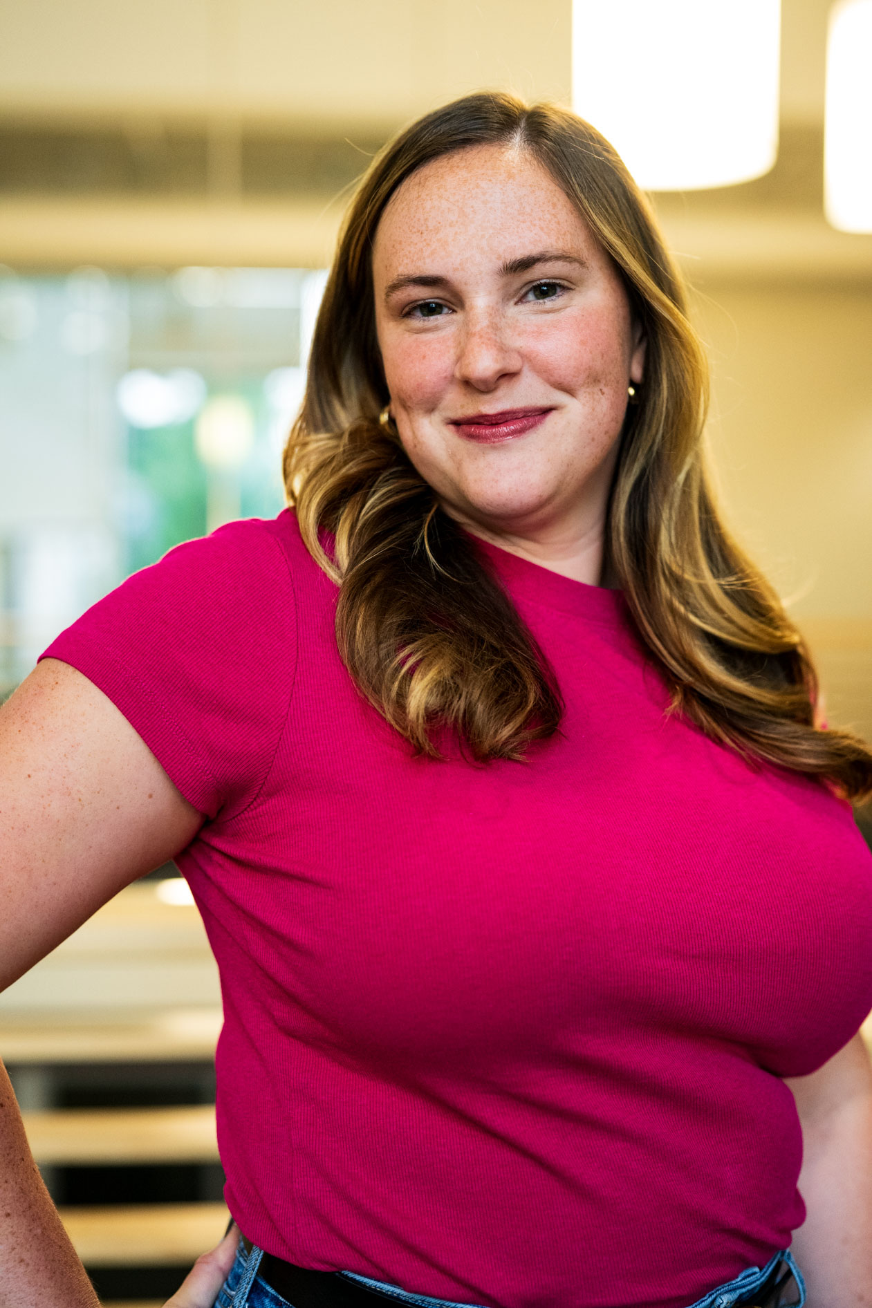 A professional headshot of a person with long light-brown hair and freckles smiling warmly. They wear a vibrant pink top against a soft-lit, blurred indoor background, capturing an approachable and authentic presence for a Planned Parenthood case study.