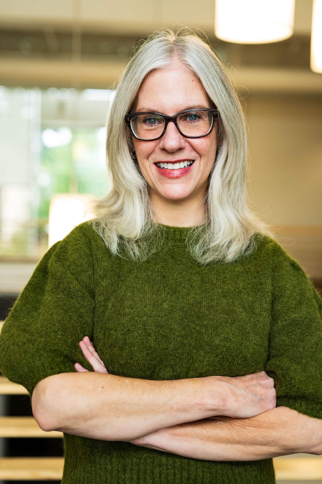 A professional portrait of a person with grey hair and glasses smiling. They wear a green textured sweater against a softly lit indoor background, capturing an approachable and confident presence.