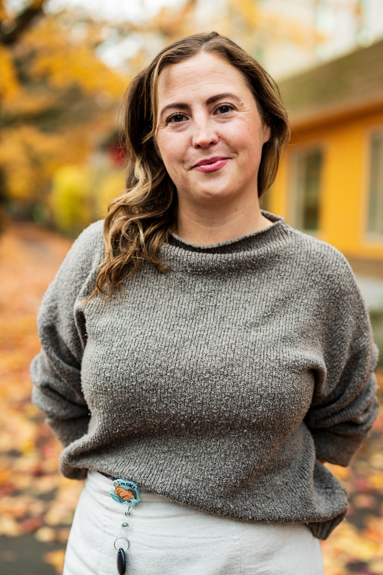 A professional outdoor portrait of a person smiling softly. They wear a grey knit sweater and a white skirt with an "I'm OK" badge reel against a blurred autumn background of yellow leaves and a building.