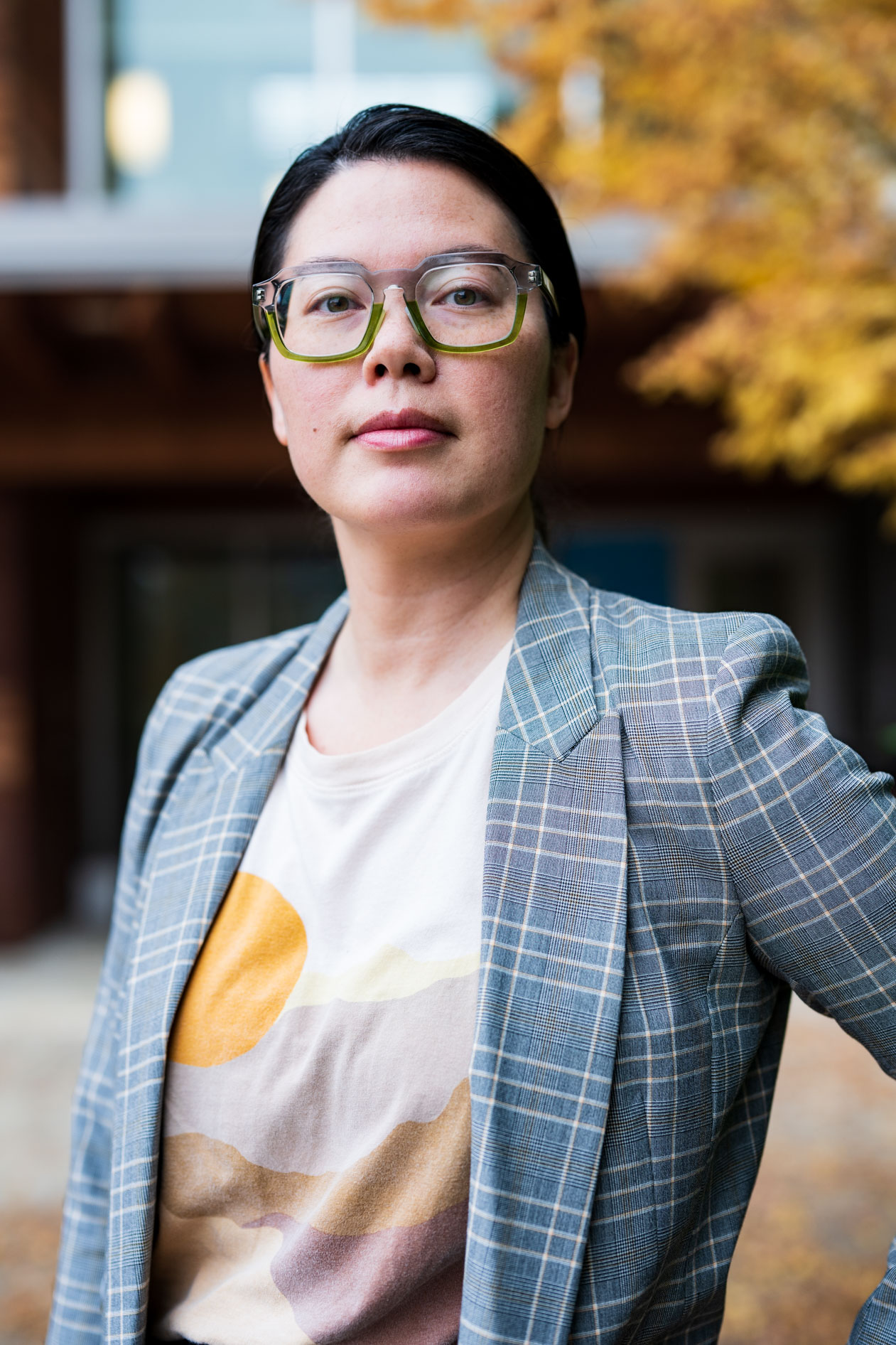A professional outdoor portrait of a person with dark hair and green-rimmed glasses. They wear a grey plaid blazer over a graphic t-shirt against a blurred autumn background, capturing a modern and authentic professional presence.