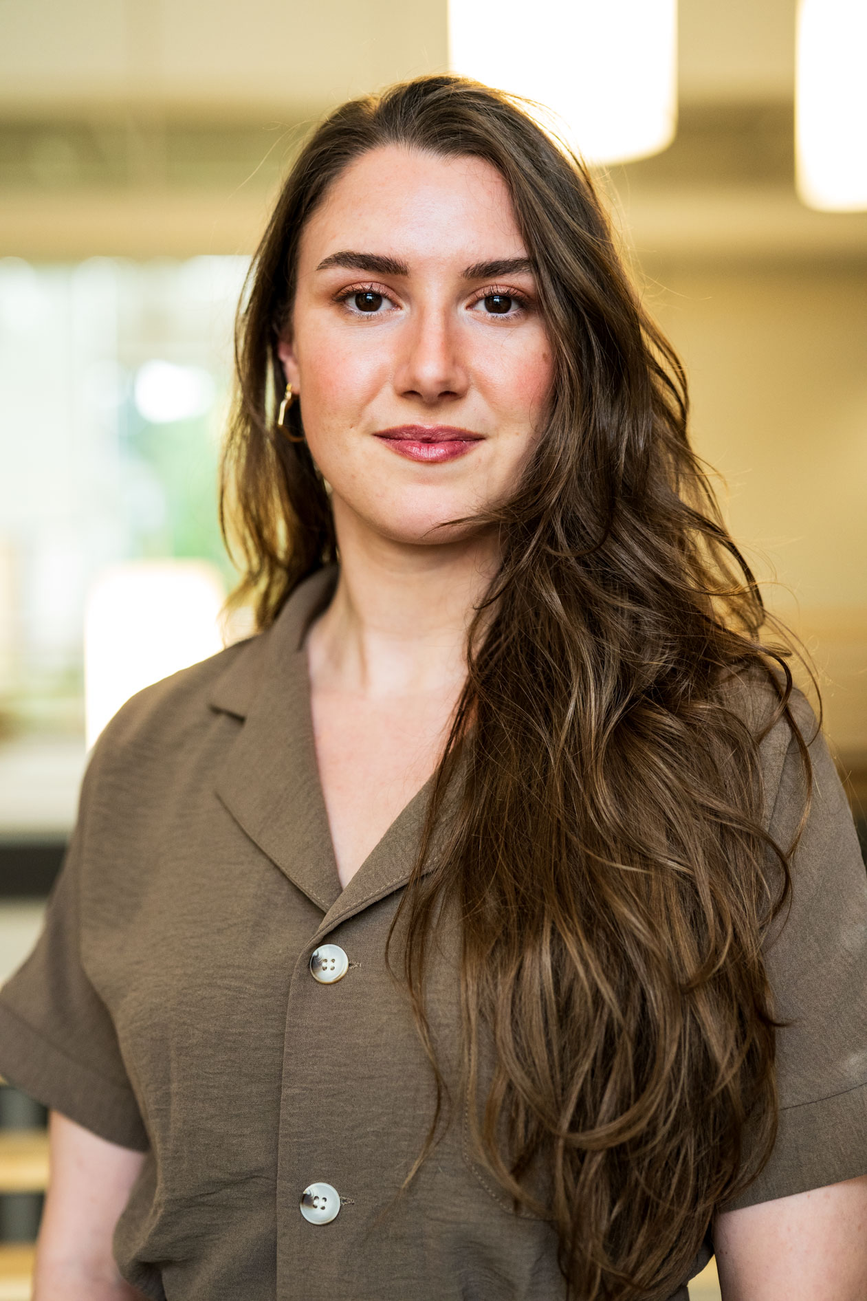 A professional headshot of a person with long, wavy brown hair and a calm expression. They wear a brown button-down shirt against a blurred, warm-toned indoor background, capturing an authentic and composed presence.