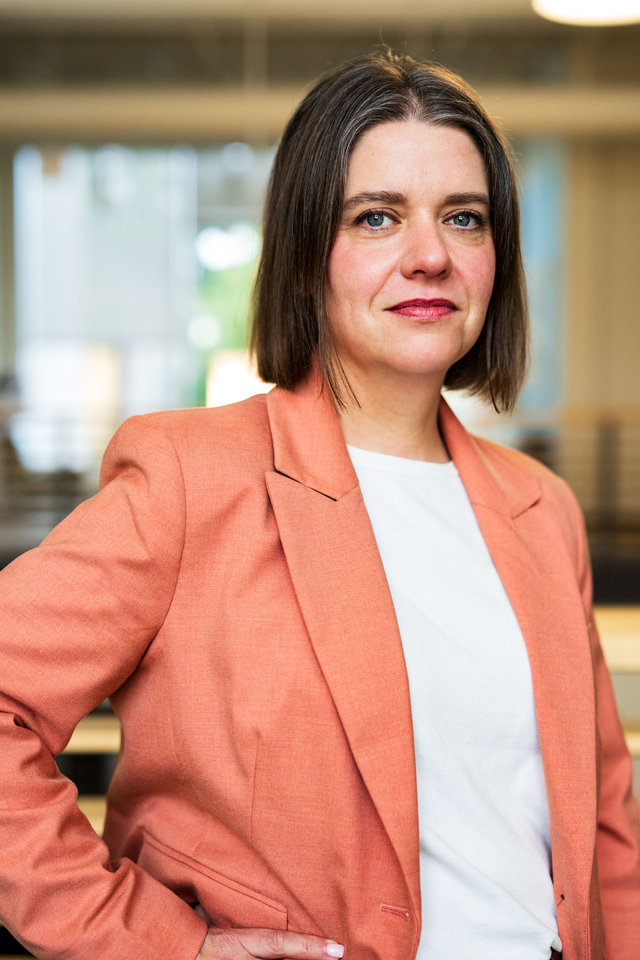 A professional headshot of a person with a brown bob hairstyle wearing a peach-colored blazer over a white top. They are standing with a hand on their hip against a blurred indoor background, capturing a poised and professional presence.