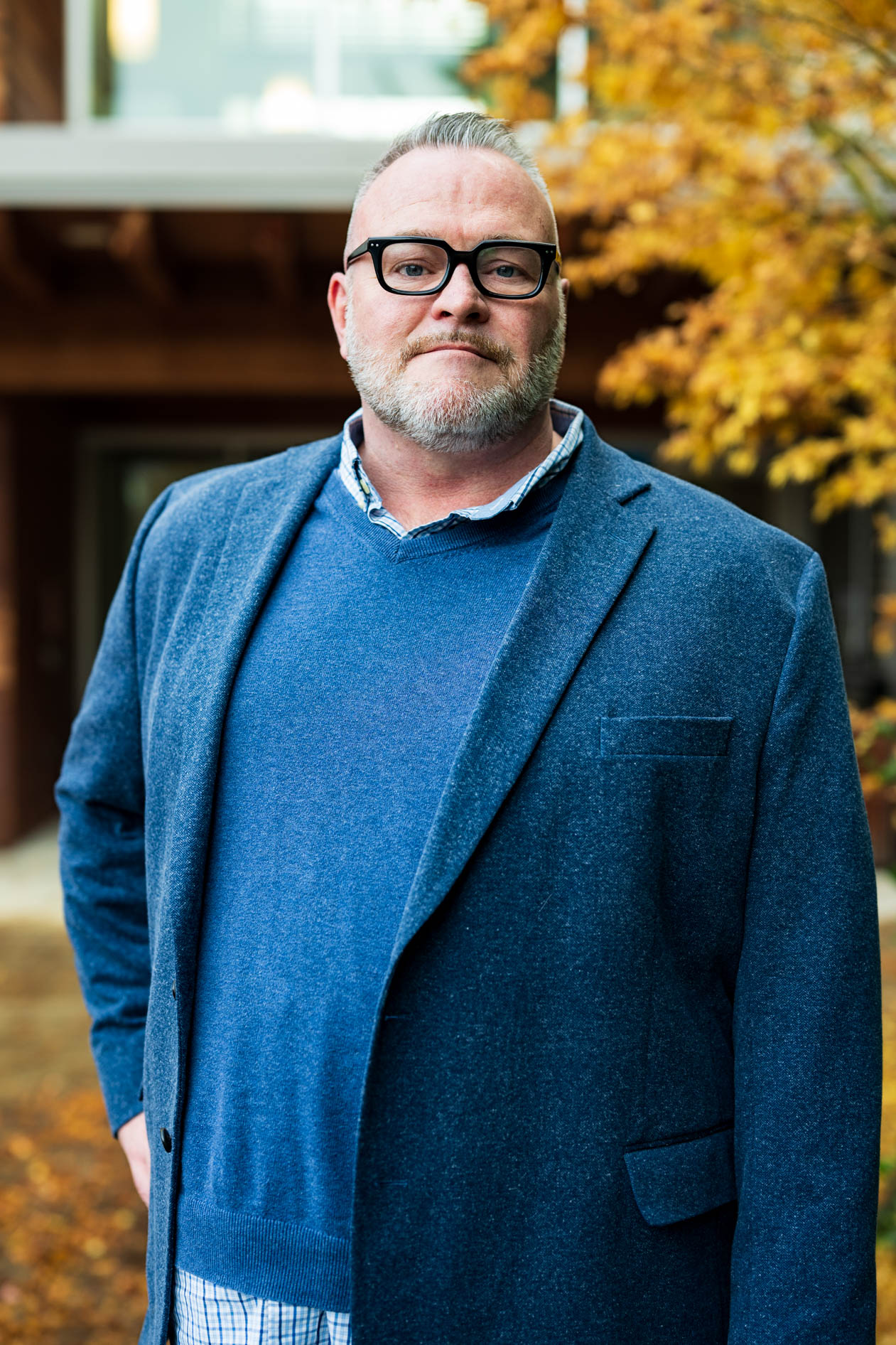 A professional outdoor portrait of a person with a beard and thick-rimmed glasses. They wear a blue blazer over a blue sweater and collared shirt against a blurred autumn background, capturing a composed and authentic professional presence.