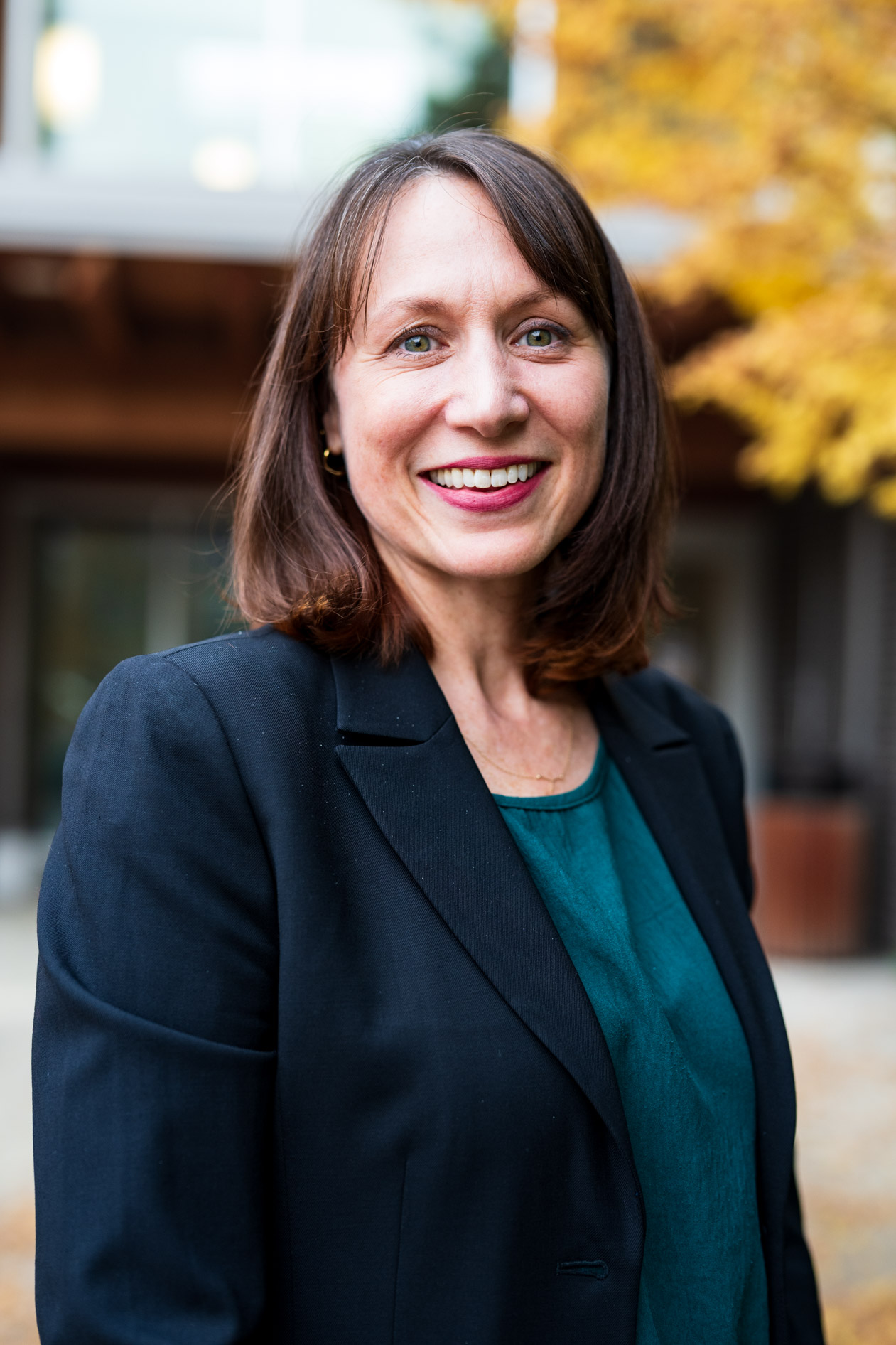 A professional outdoor portrait of a person with a brown bob hairstyle smiling warmly. They wear a black blazer over a dark green top against a blurred autumn background, capturing a friendly and authentic professional presence.
