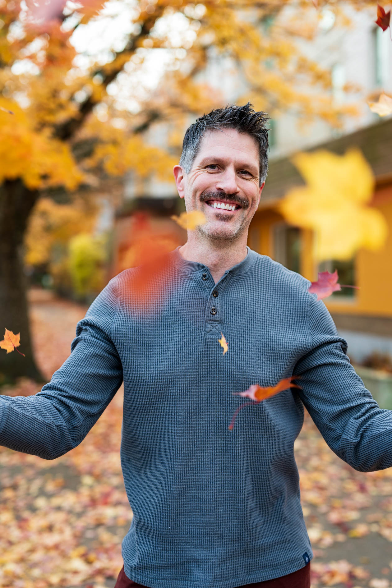 A person with a mustache smiles joyfully as autumn leaves fall around them. They wear a grey henley shirt against a blurred background of yellow fall foliage, capturing a candid and energetic moment for a Planned Parenthood headshot session.
