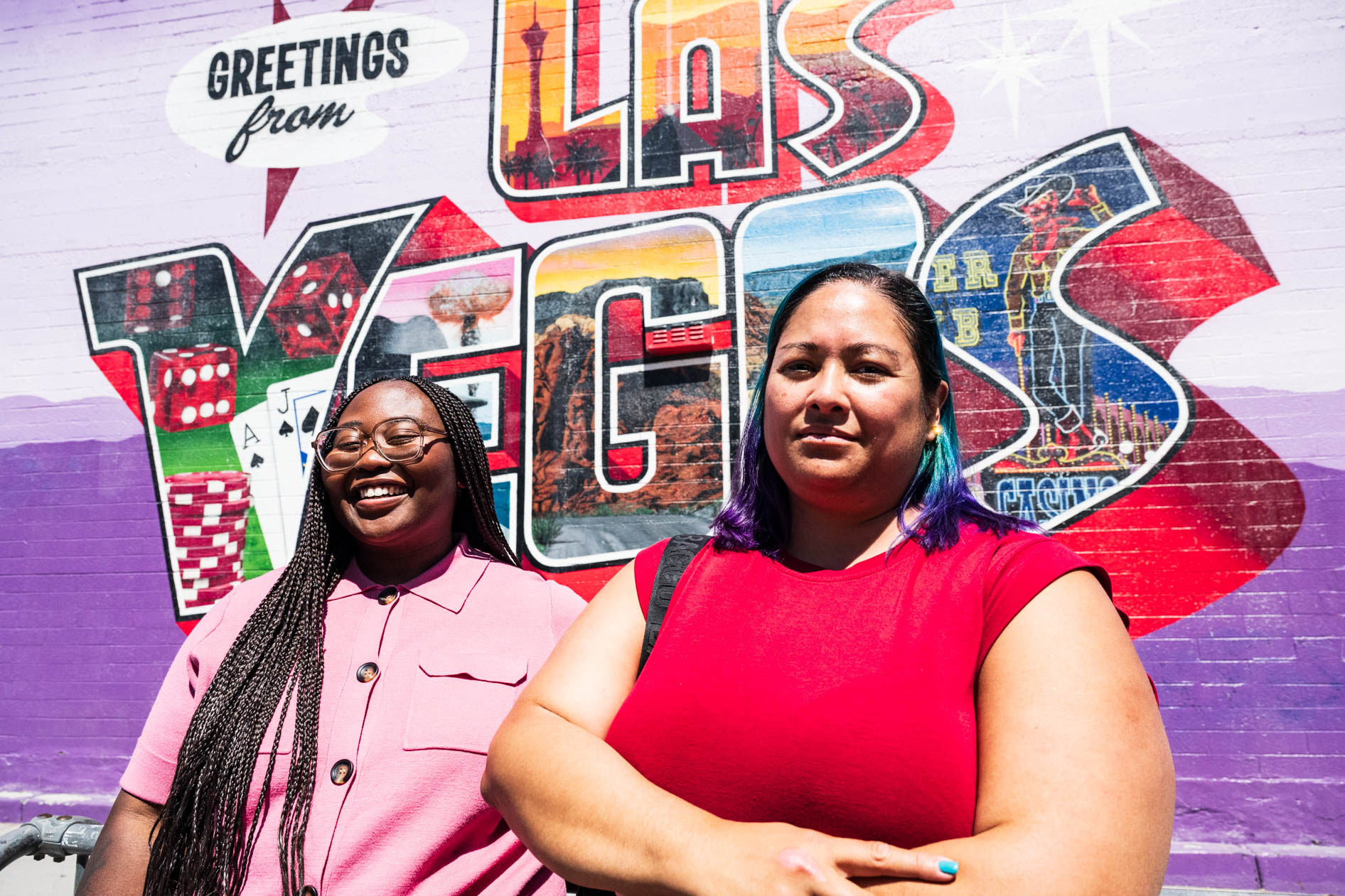 Two people pose outdoors in front of a colorful "Greetings from Las Vegas" postcard-style mural. One person laughs joyfully in a pink collared shirt, while the other stands confidently with arms crossed in a red top, celebrating the marketing agency's expansion.