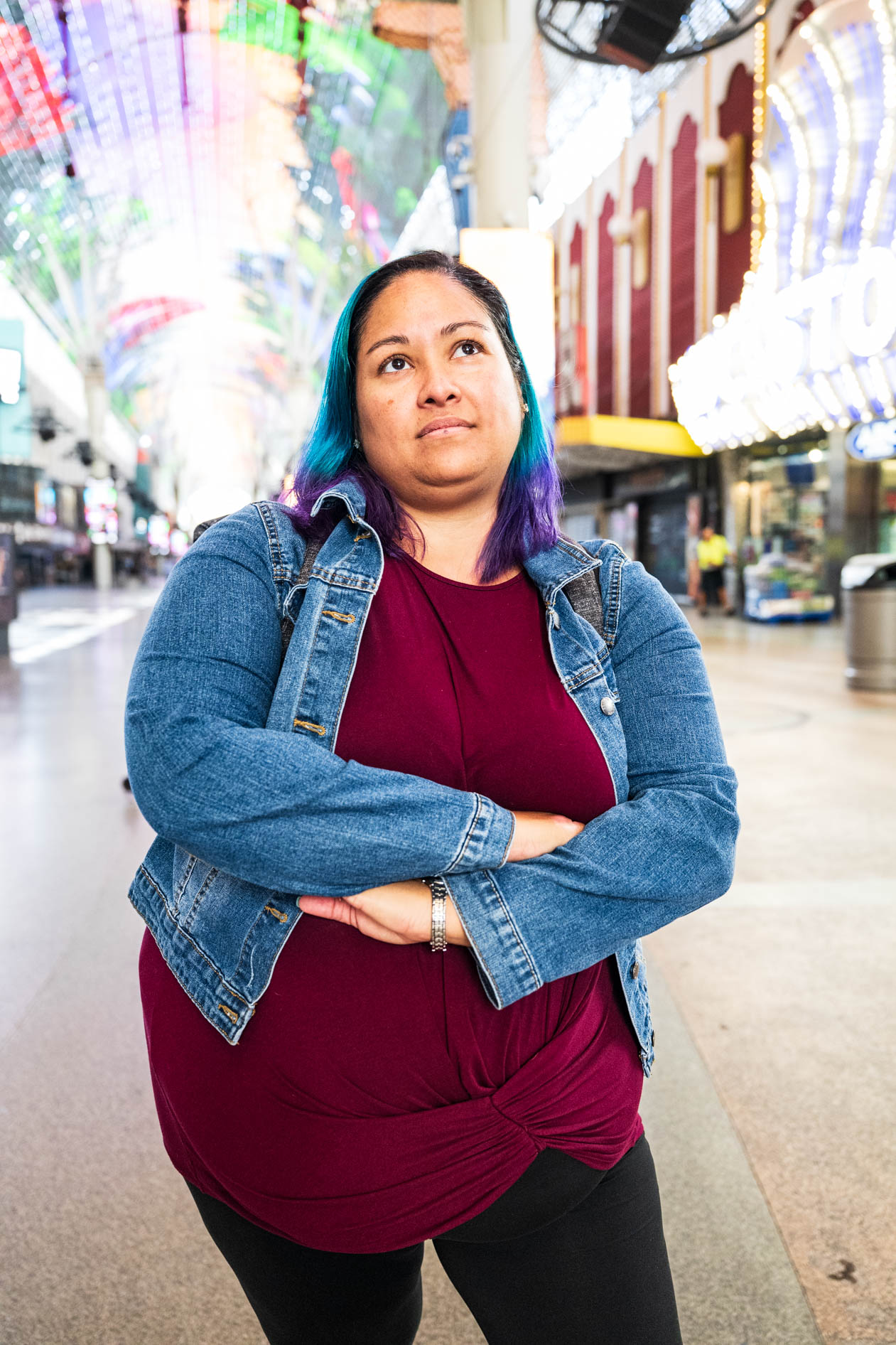 A portrait of a person with blue and purple hair standing on Fremont Street in Las Vegas with arms crossed. They look upward thoughtfully, wearing a denim jacket over a maroon top under the prominent light canopy.