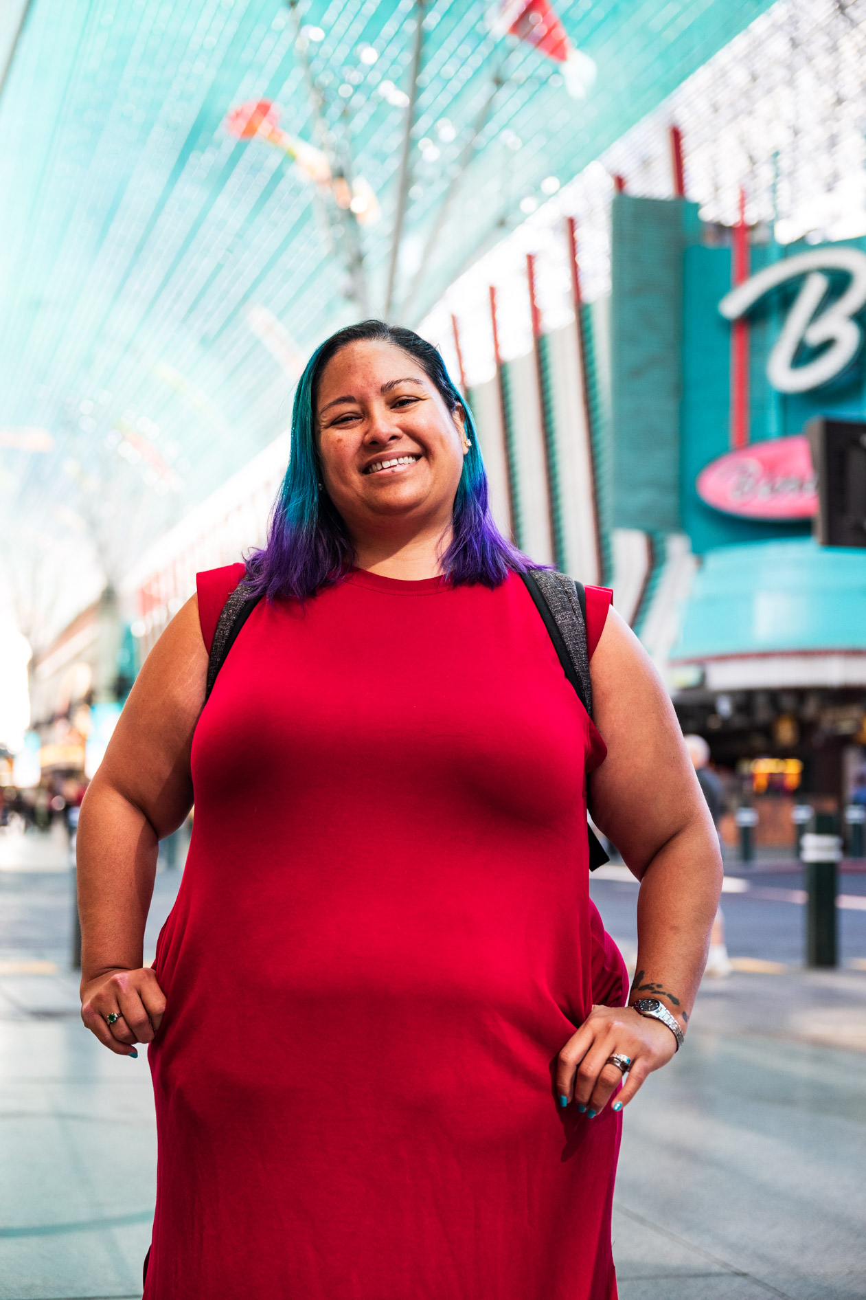 A portrait of a person with blue and purple hair smiling warmly on Fremont Street in Las Vegas. They wear a red dress and a backpack, posing with hands on hips under the prominent light canopy for an Ashbean PDX agency expansion photoshoot.