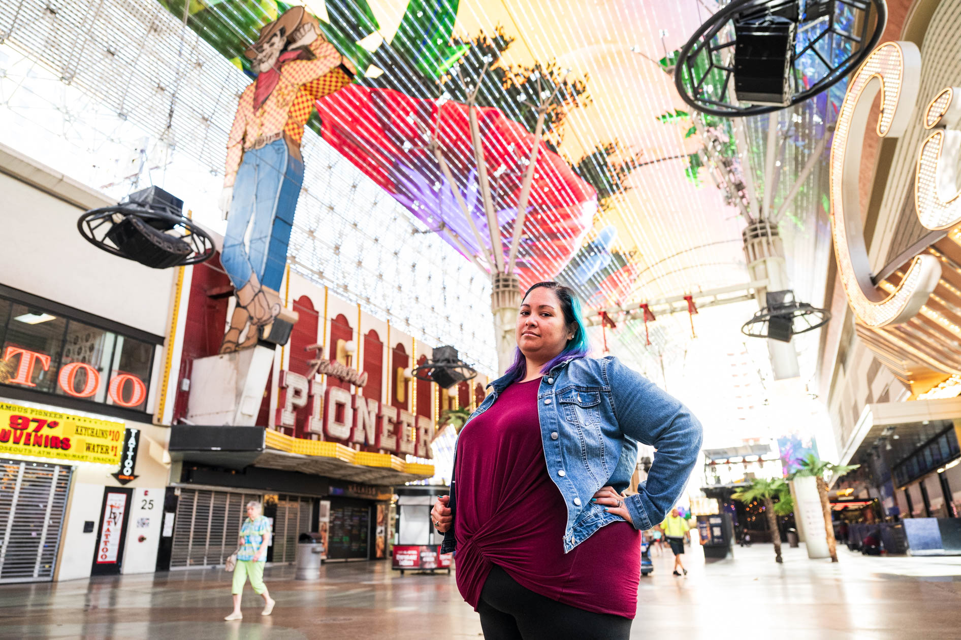 A person with blue and purple hair stands confidently with a hand on their hip on Fremont Street in Las Vegas. They wear a denim jacket over a maroon top, with the historic Vegas Vic cowboy neon sign and Pioneer club visible in the background under the lig
