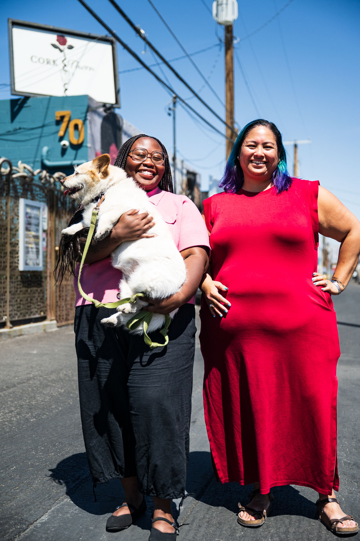 Two people smile brightly while standing outdoors on a sunny street. One person in a pink shirt cradles a small white dog, and the other stands in a long red dress. A sign reading "Cork & Thorn" is visible on the building behind them.