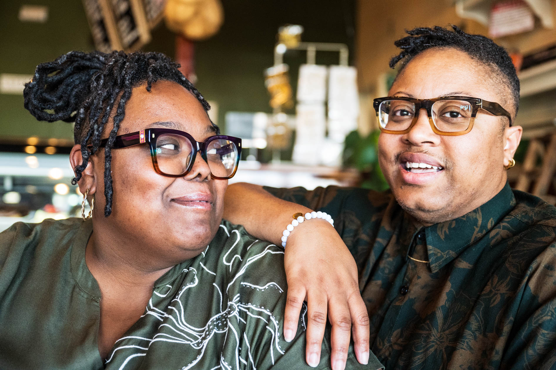 A close-up portrait of two people wearing glasses and earth-toned patterned shirts. They are smiling and leaning toward each other in a brightly lit indoor space, capturing a sense of connection during a commercial photoshoot.