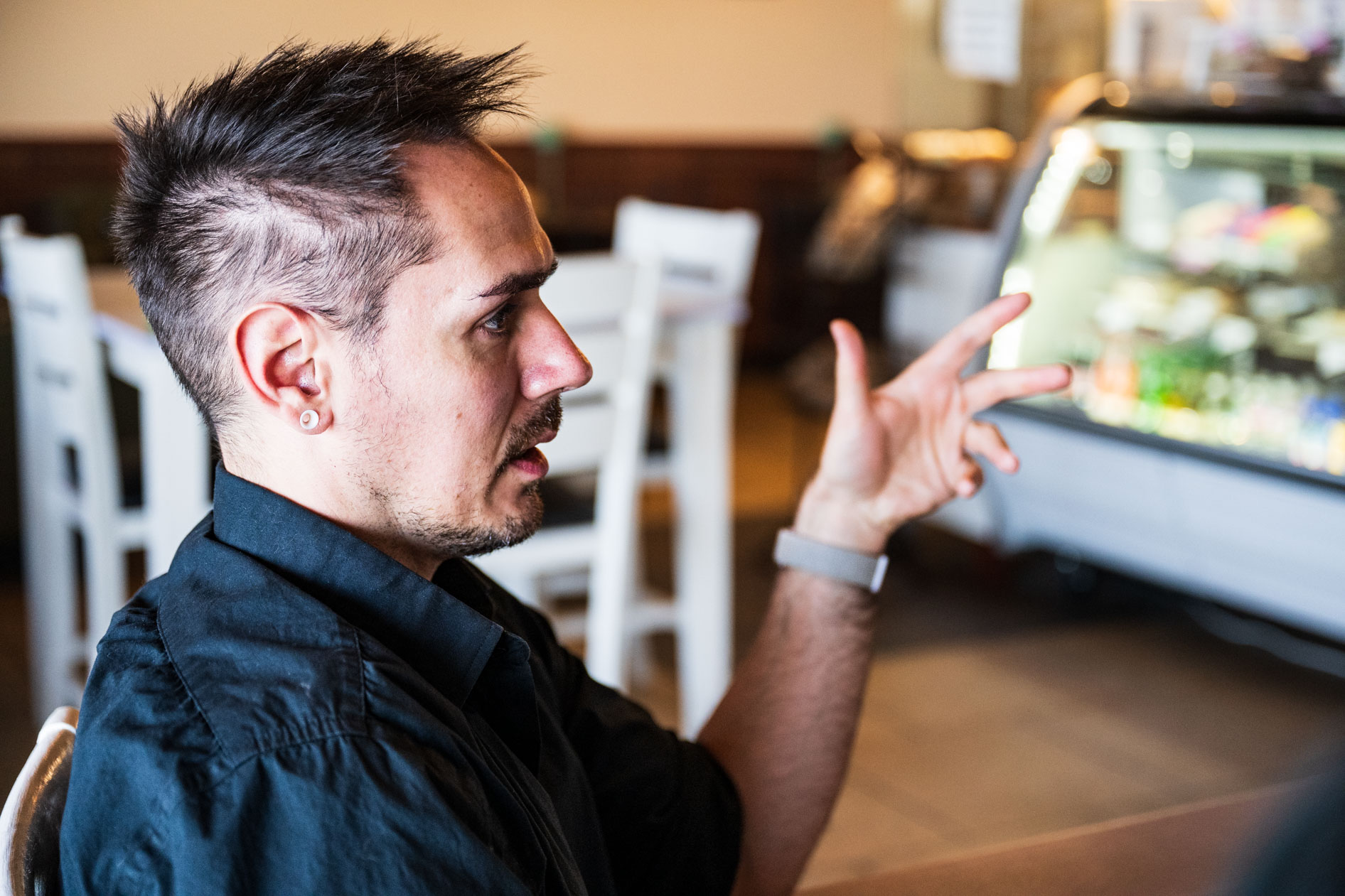 A side profile of a person with styled dark hair and an ear gauge talking in a cafe. They are gesturing with their hand while wearing a black button-down shirt, with blurry tables and a display case behind them.