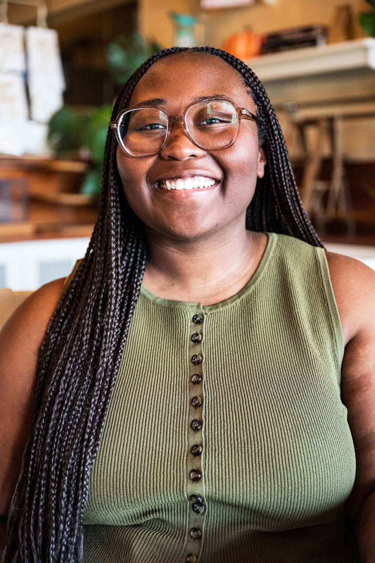 A professional headshot of a person with long braided hair and glasses smiling warmly. They wear an olive green sleeveless button-front top in a brightly lit indoor space with a blurred background.