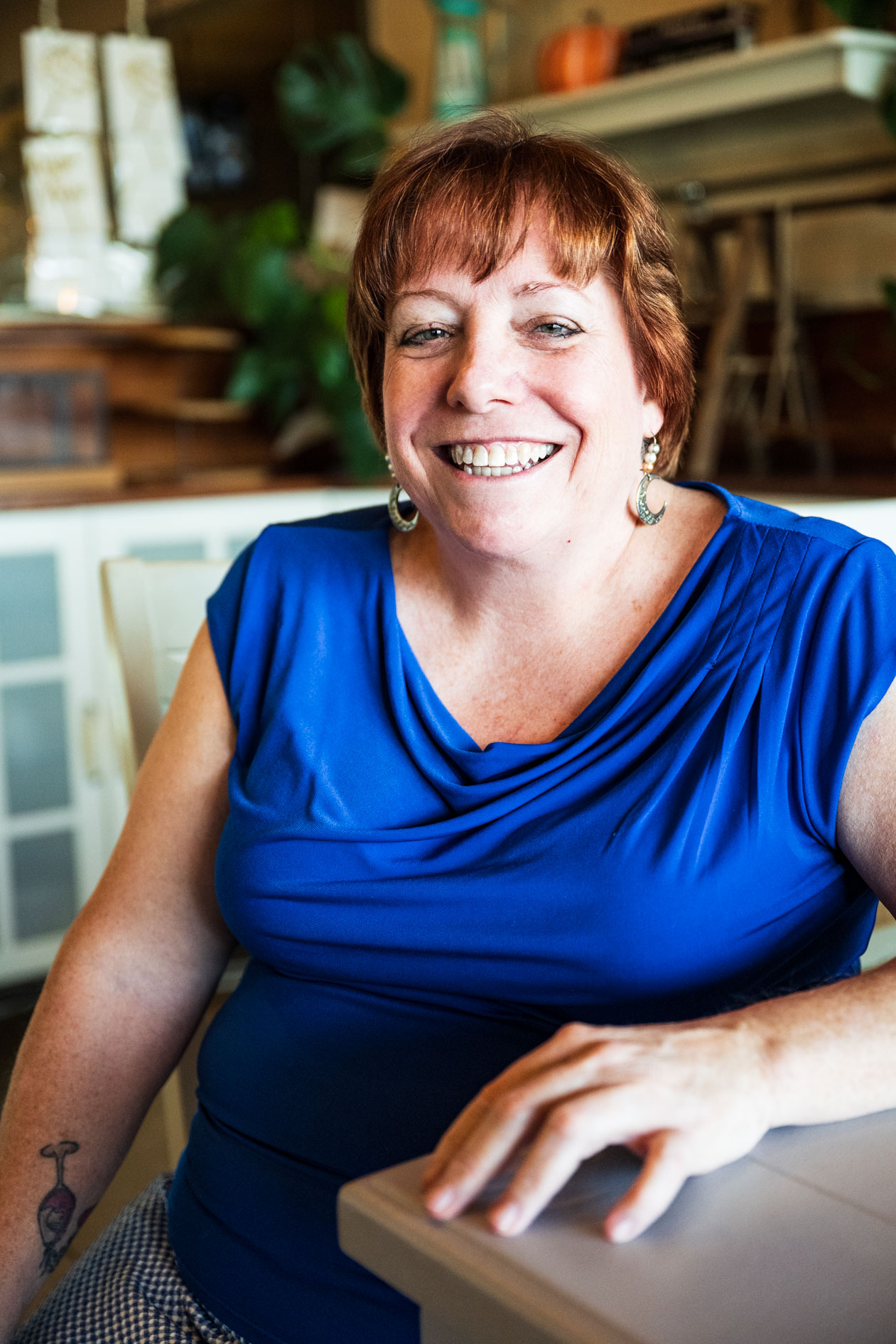 A professional portrait of a smiling person with short red hair sitting at a table. They wear a vibrant blue top and crescent moon earrings in a brightly lit indoor space with a blurred background.