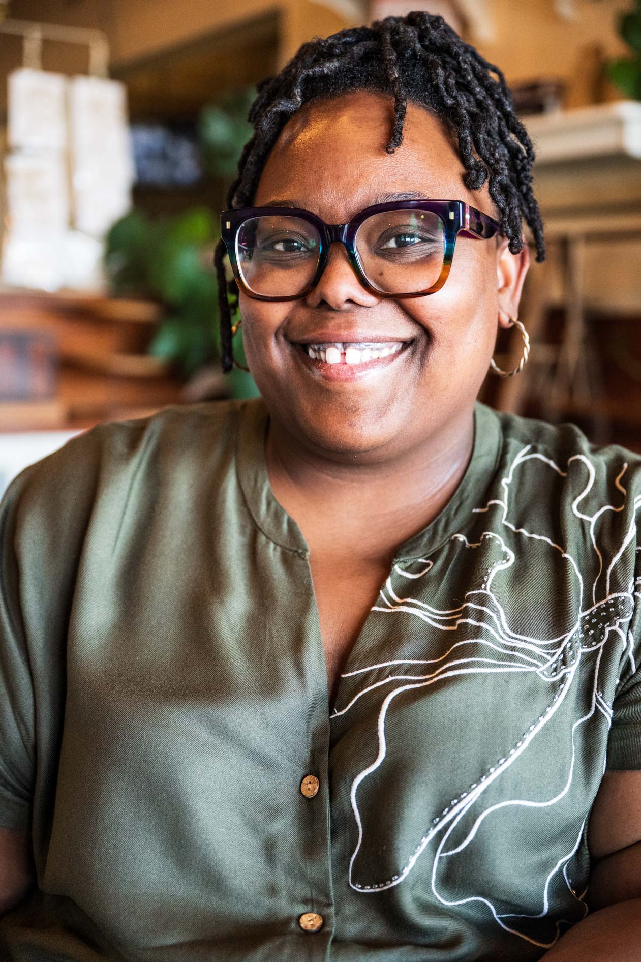 A professional headshot of a smiling person with short locs and glasses. They wear an olive green shirt with a white line-art floral pattern in a brightly lit indoor space with a blurred background.