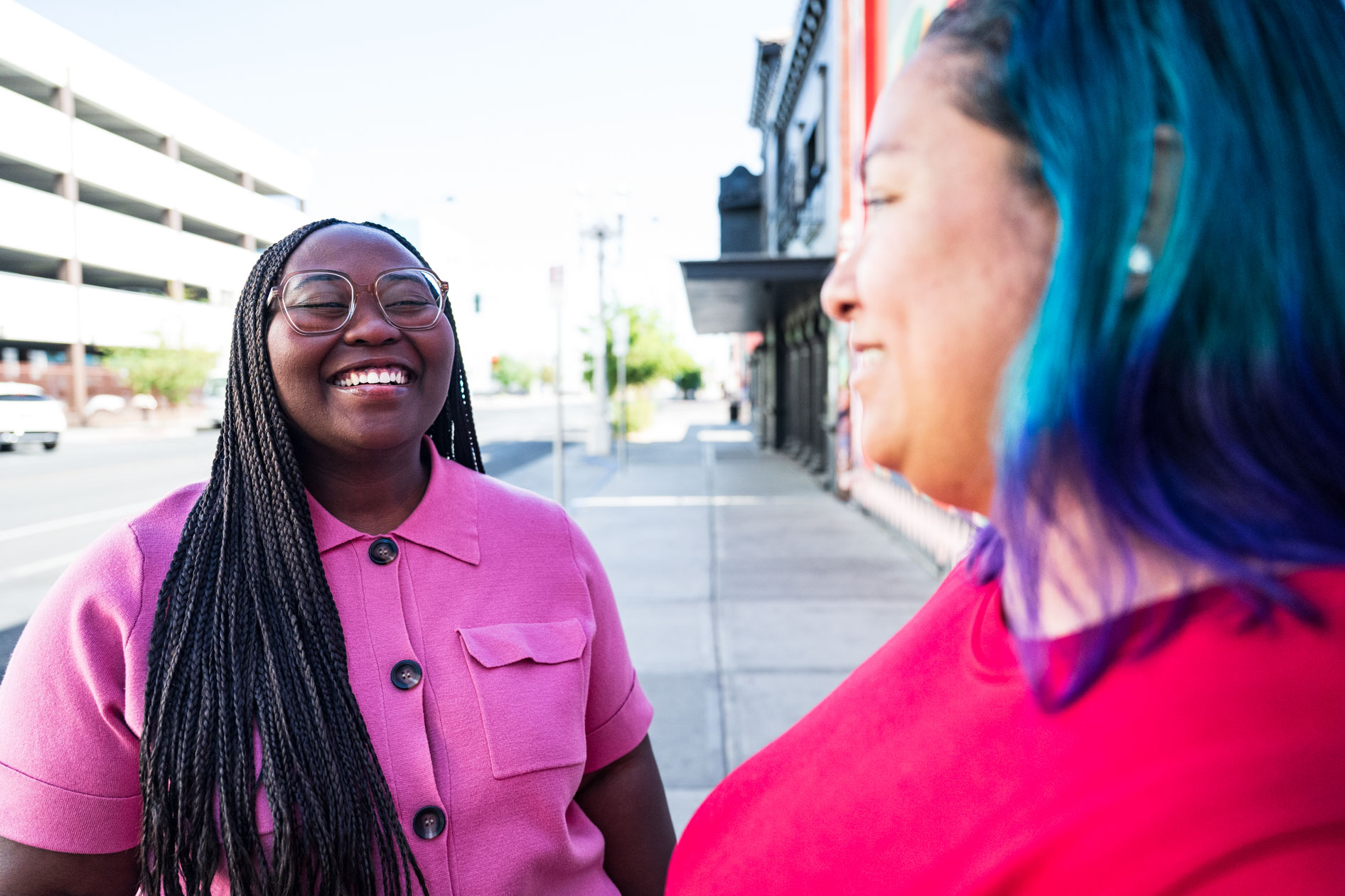 A candid moment from a commercial photoshoot showing a person with long braids and glasses laughing joyfully while talking to a person with blue and purple hair. They are standing on a sunny city sidewalk in Las Vegas for the Ashbean PDX agency expansion.