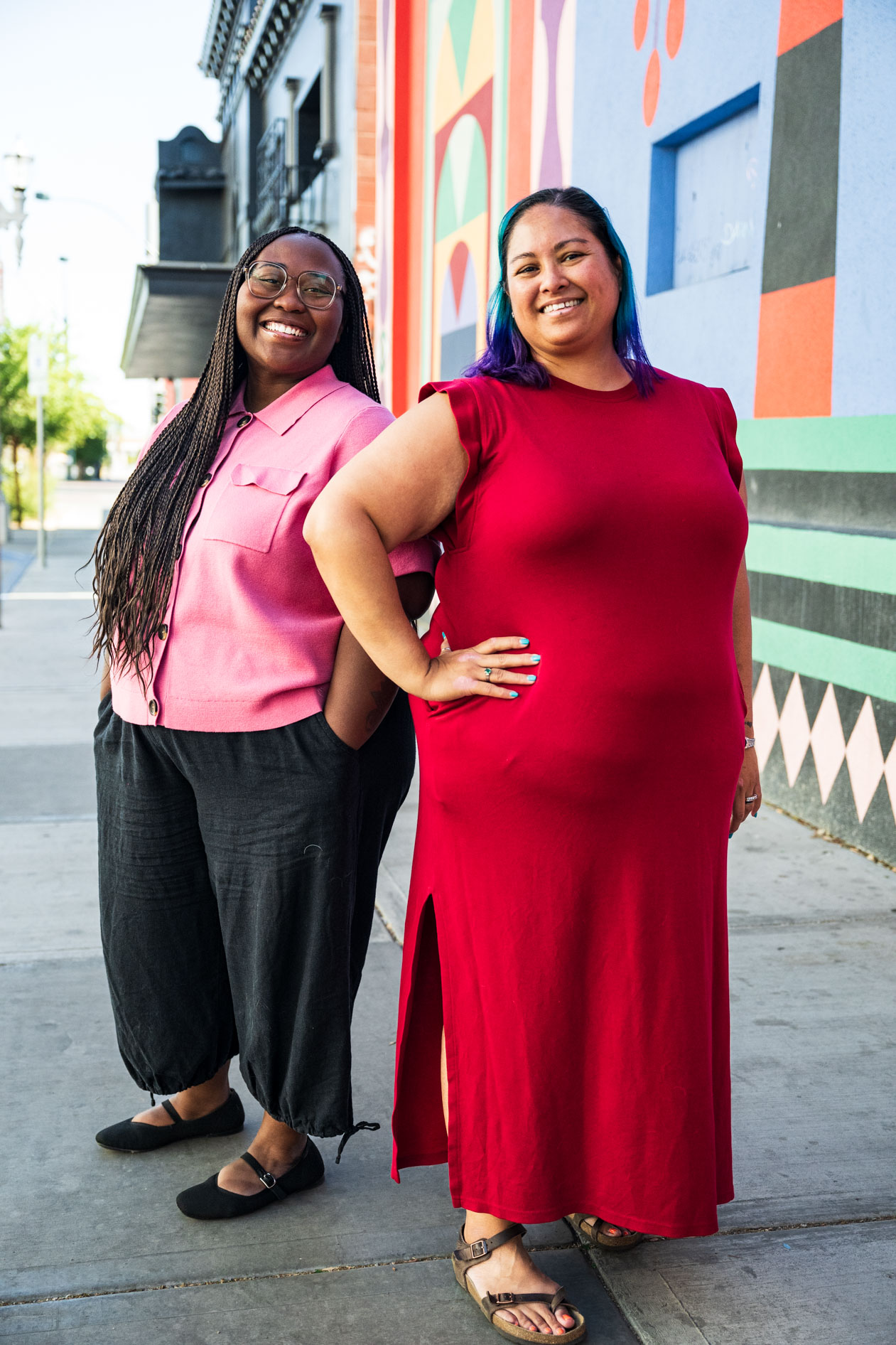 Two people smile brightly while standing outdoors on a city sidewalk next to a brightly patterned, colorful wall. One person wears a pink shirt with black pants, and the other stands with a hand on their hip in a long red dress.