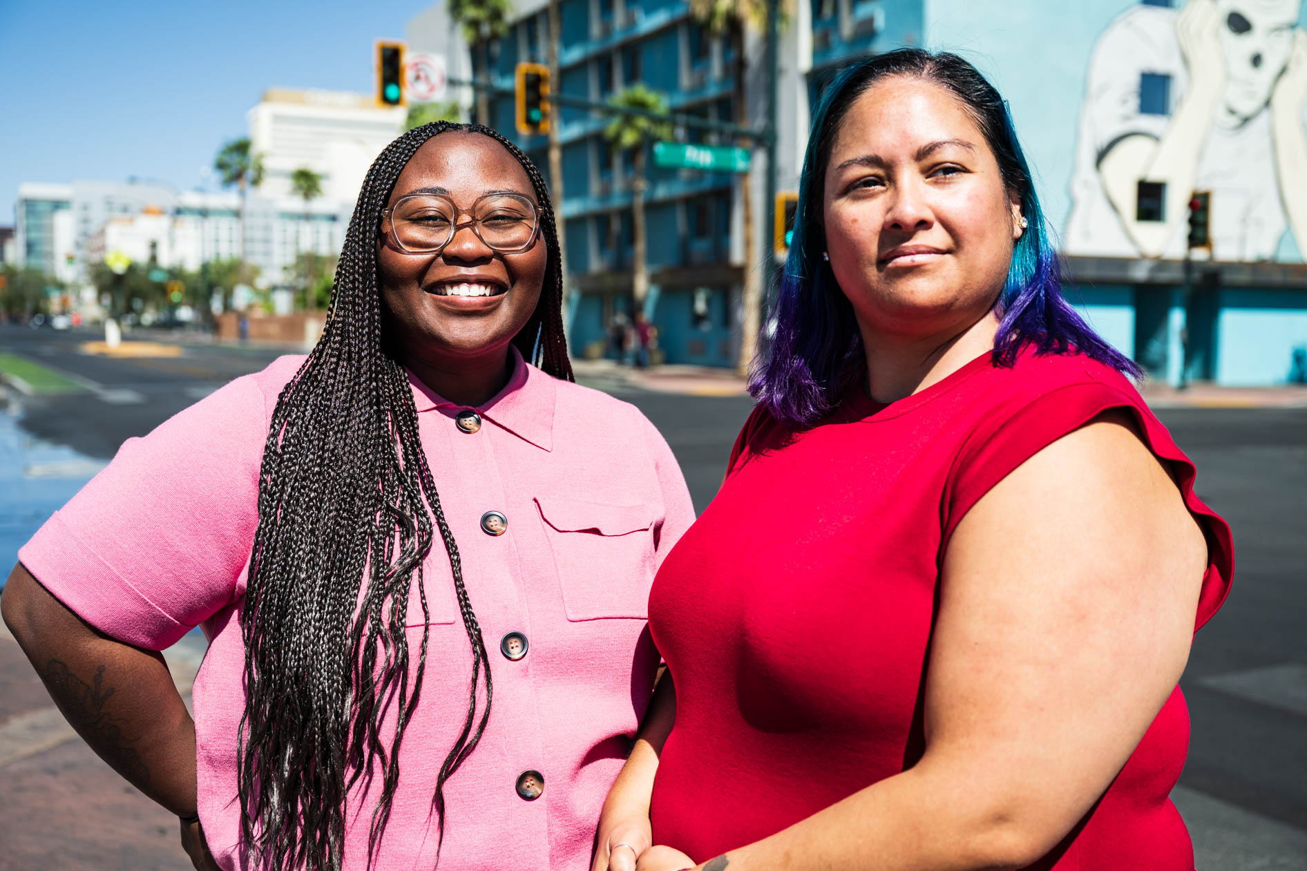 Two people smile while standing outdoors on a sunny city street. One person wears a pink collared shirt, and the other wears a red top, with city buildings and a mural visible in the background.