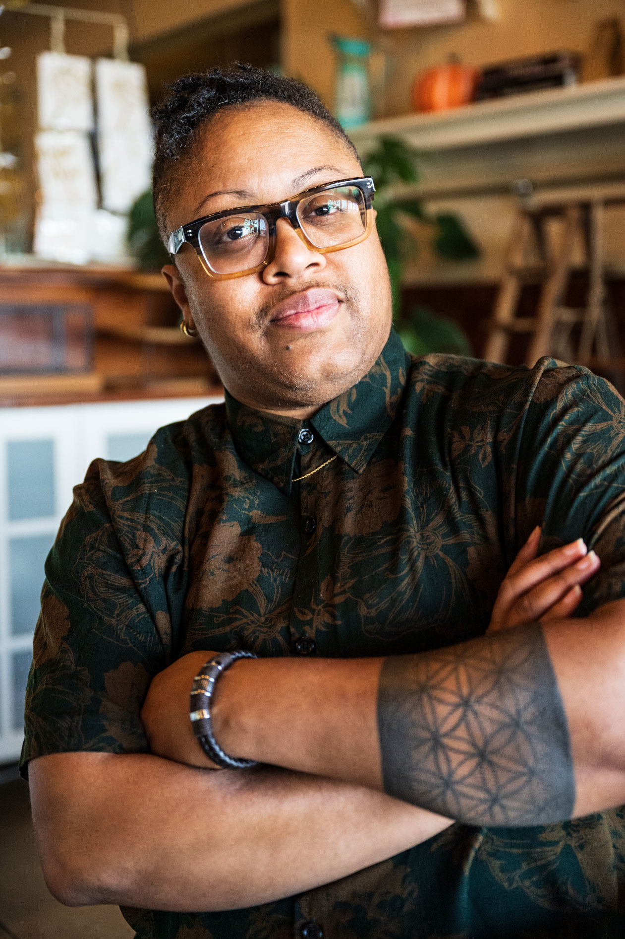 A professional headshot of a person with short hair and glasses standing with arms crossed. They wear a dark, earth-toned floral button-down shirt and have a geometric forearm tattoo in a brightly lit indoor space.