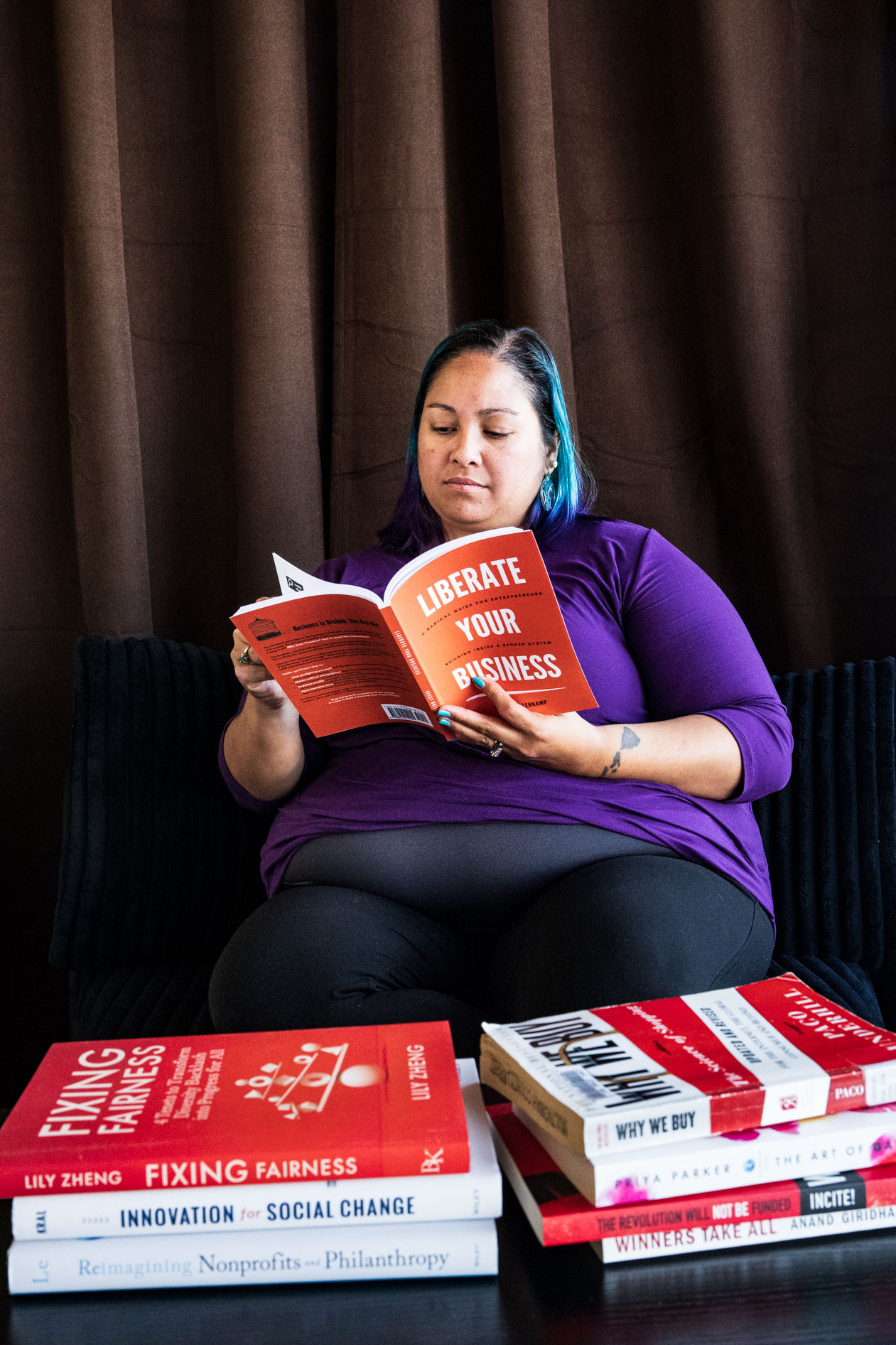 A person with blue and purple hair sits on a chair reading a book titled "Liberate Your Business." They wear a purple long-sleeve shirt, with several other business and social impact books stacked on a table in the foreground.