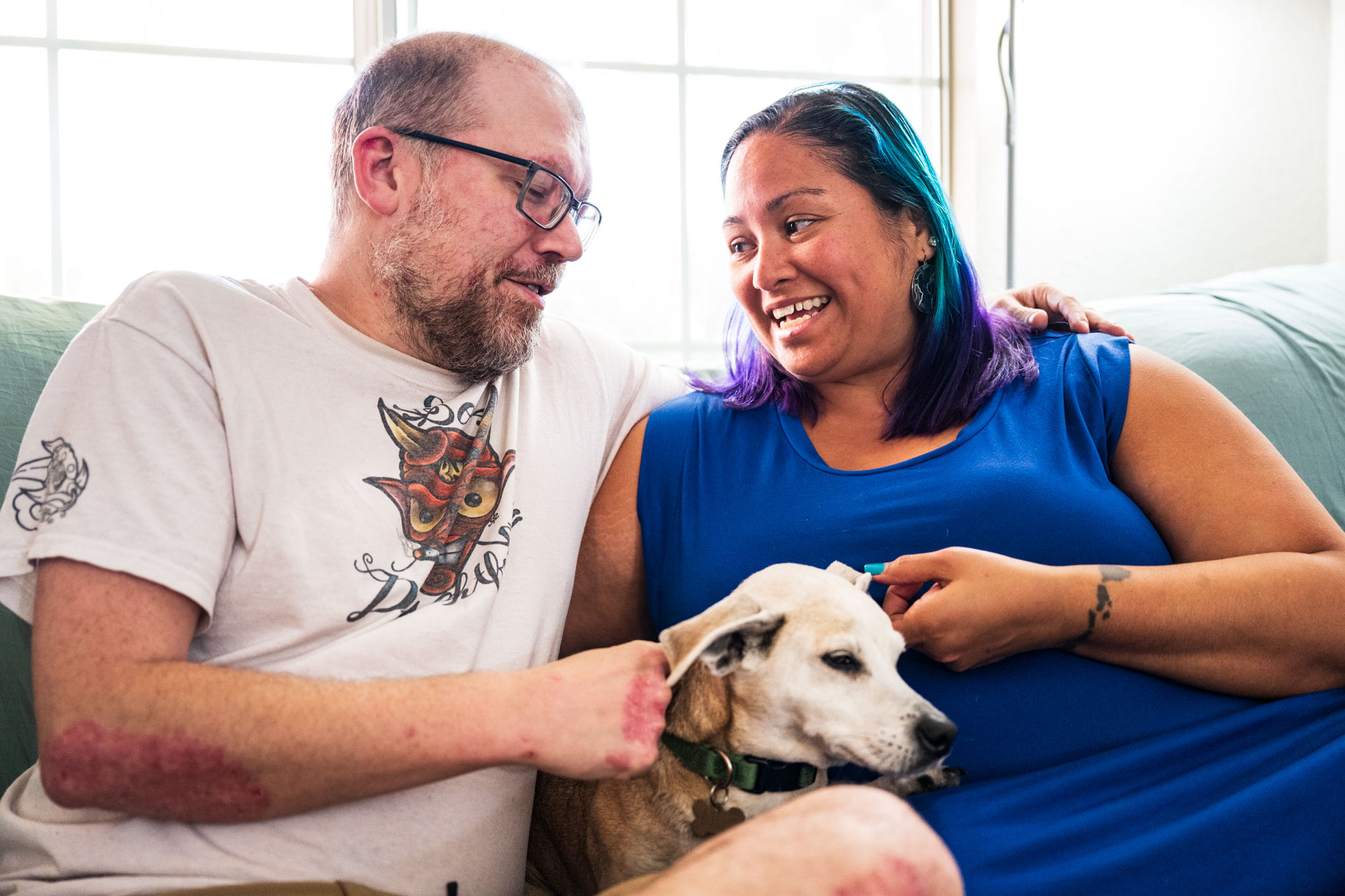 Two people sit closely on a couch smiling and looking at each other while petting a small dog. One person wears glasses and a graphic t-shirt, and the other wears a blue dress and has vibrant purple and blue hair.