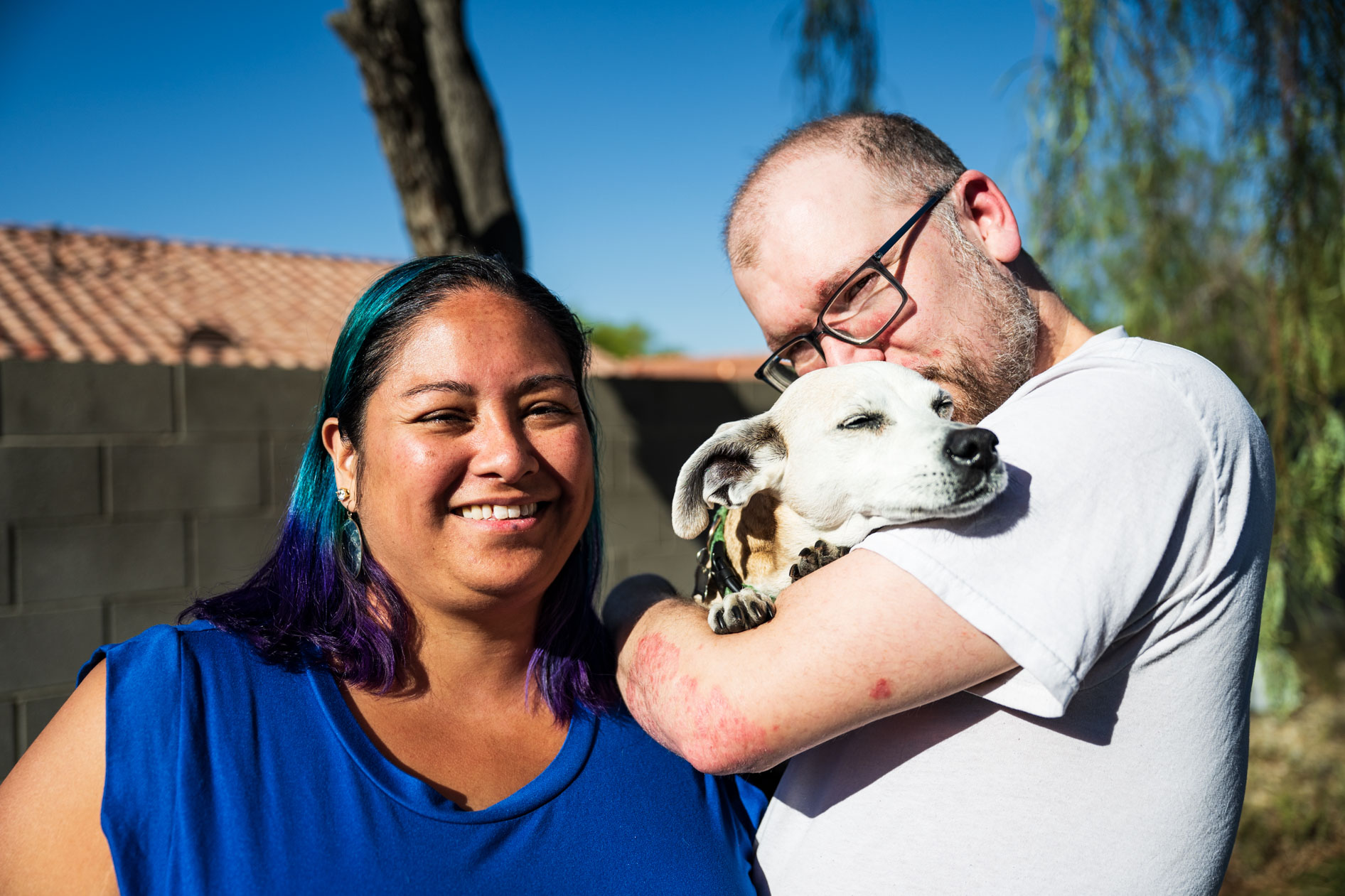 Two people pose outdoors on a sunny day. One person with blue and purple hair smiles at the camera, while the other person cradles a small dog in their arms against a backdrop of a brick wall and clear blue sky.