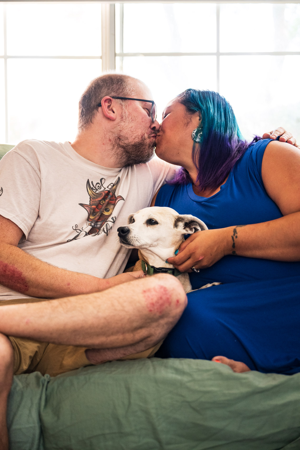Two people share a kiss while sitting together on a couch with a small dog between them. One person wears a graphic t-shirt and glasses, while the other has vibrant blue and purple hair and wears a bright blue dress.