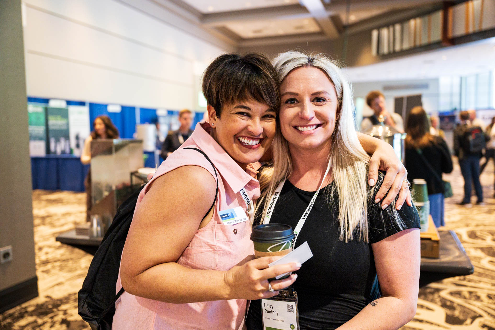 Two women smile and embrace warmly inside a convention center hall at the Efficiency Exchange 2026 conference. They wear event lanyards and name tags, with trade show booths and other attendees blurred in the background.