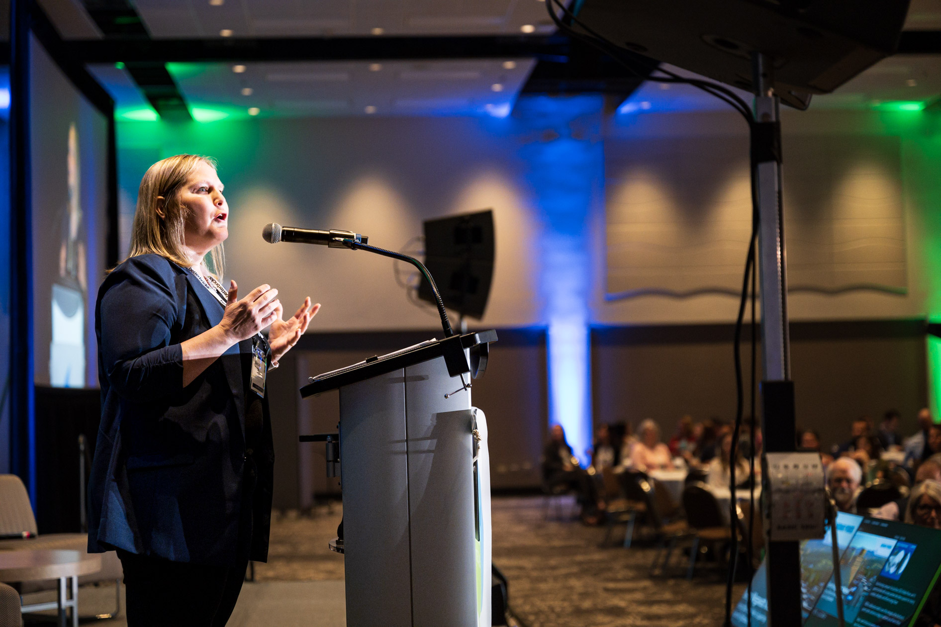 A speaker stands at a podium addressing an audience inside a large auditorium at the Efficiency Exchange 2026 conference. They wear a dark blazer and gesture while speaking into a microphone, with blue and green event lighting illuminating the background.
