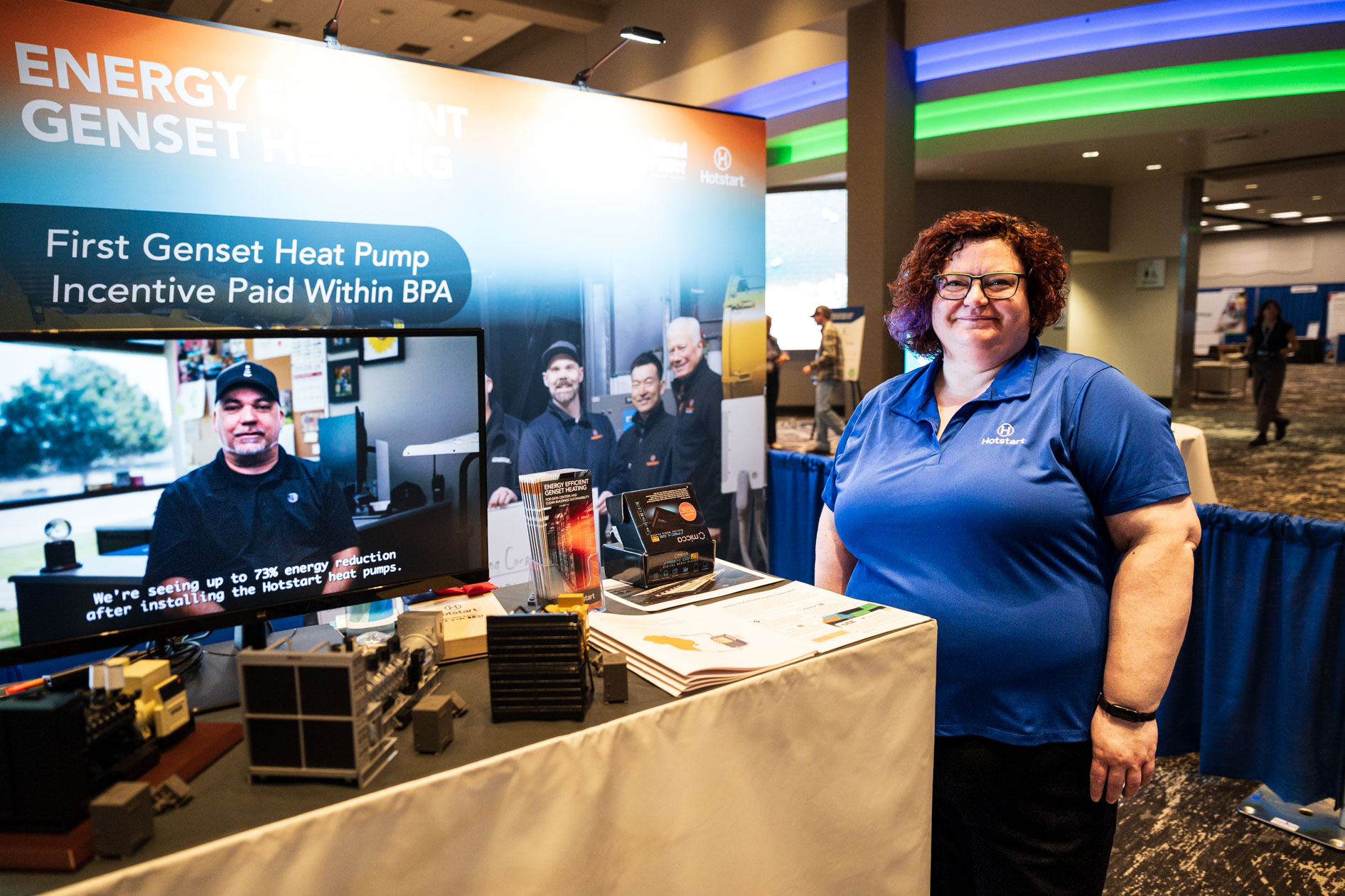 An exhibitor in a blue polo stands by their Hotstart booth at the Efficiency Exchange 2026 conference. The display includes a video monitor showcasing a testimonial alongside product models on the table.