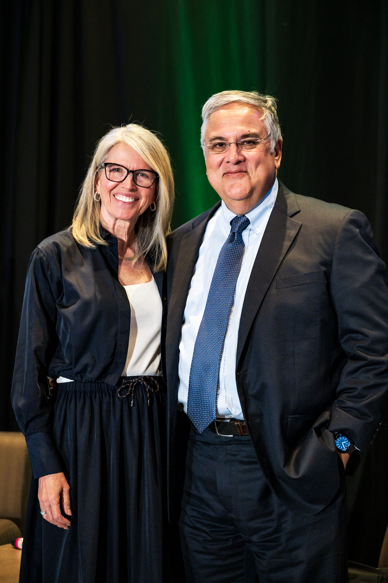 Two people pose together smiling at the Efficiency Exchange 2026 conference. One wears a black dress with glasses, and the other wears a dark suit with a blue tie against a dark green backdrop.