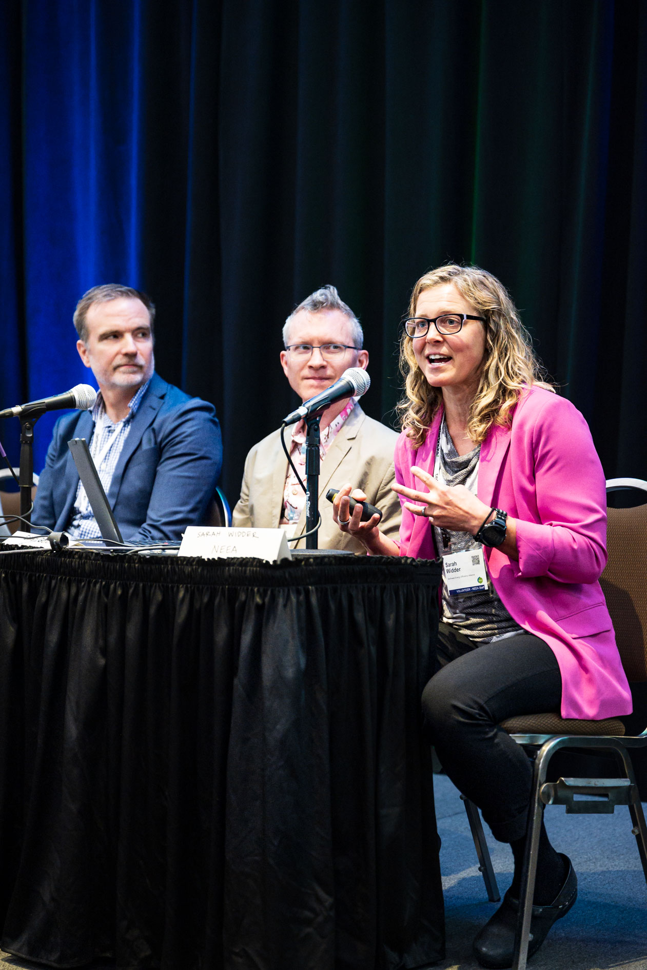 Three panelists sit at a table during a presentation at the Efficiency Exchange 2026 conference. A woman in a pink blazer speaks into a microphone while gesturing, as two other male panelists listen beside her against a dark curtain backdrop.