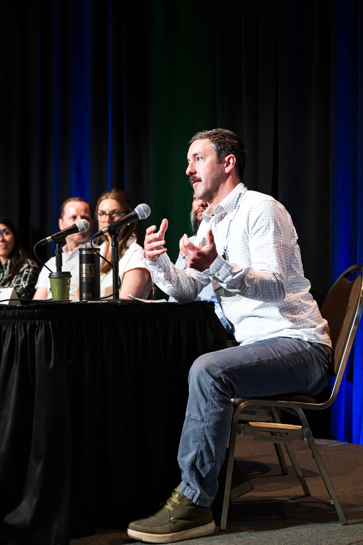 A panelist in a checkered shirt speaks while gesturing at a table during the Efficiency Exchange 2026 conference. Other panelists listen in the background on stage against a dark curtain.