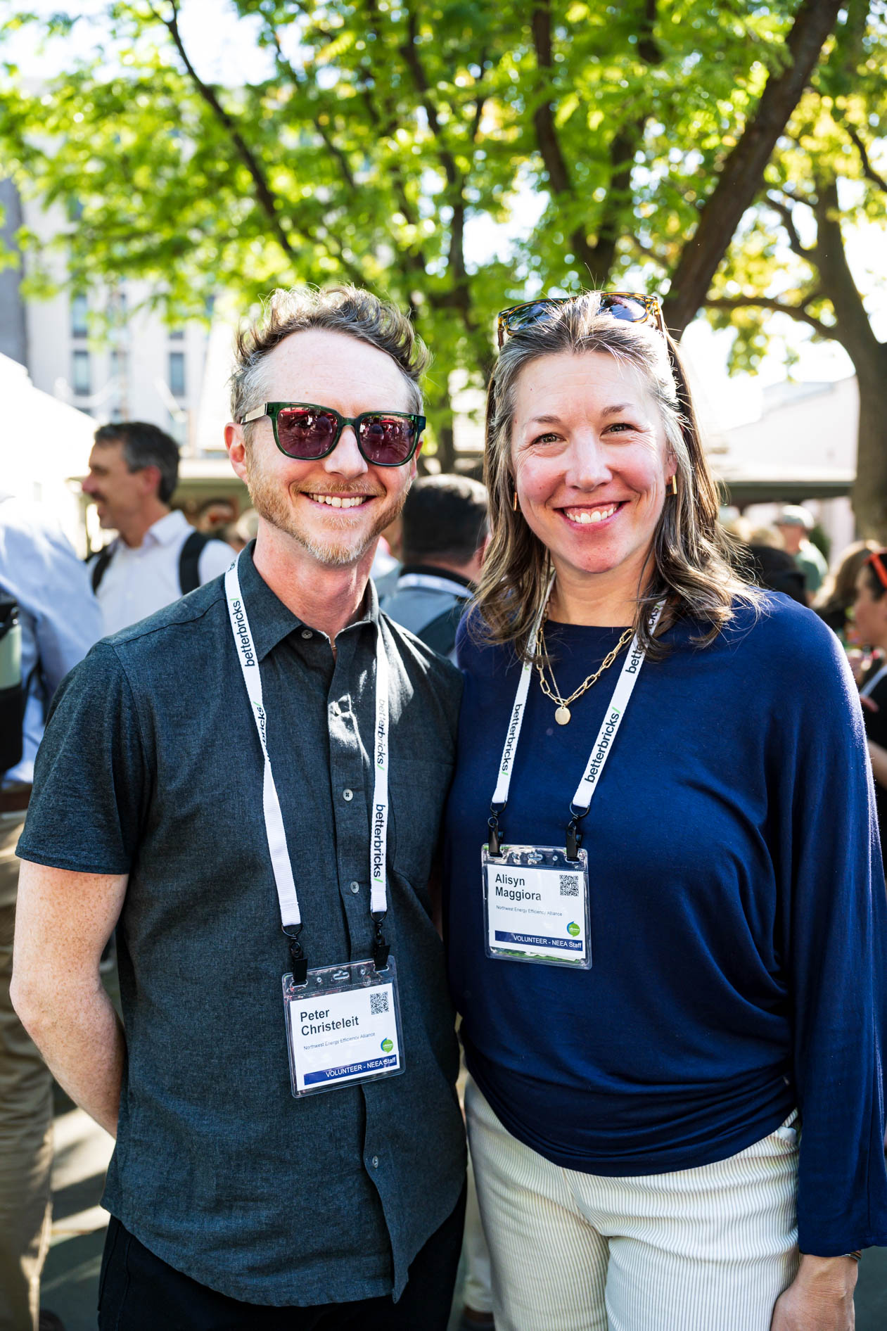 Two attendees smile outdoors at the Efficiency Exchange 2026 conference. One wears a dark short-sleeve button-down shirt with sunglasses, and the other wears a blue long-sleeve top, both displaying event lanyards.