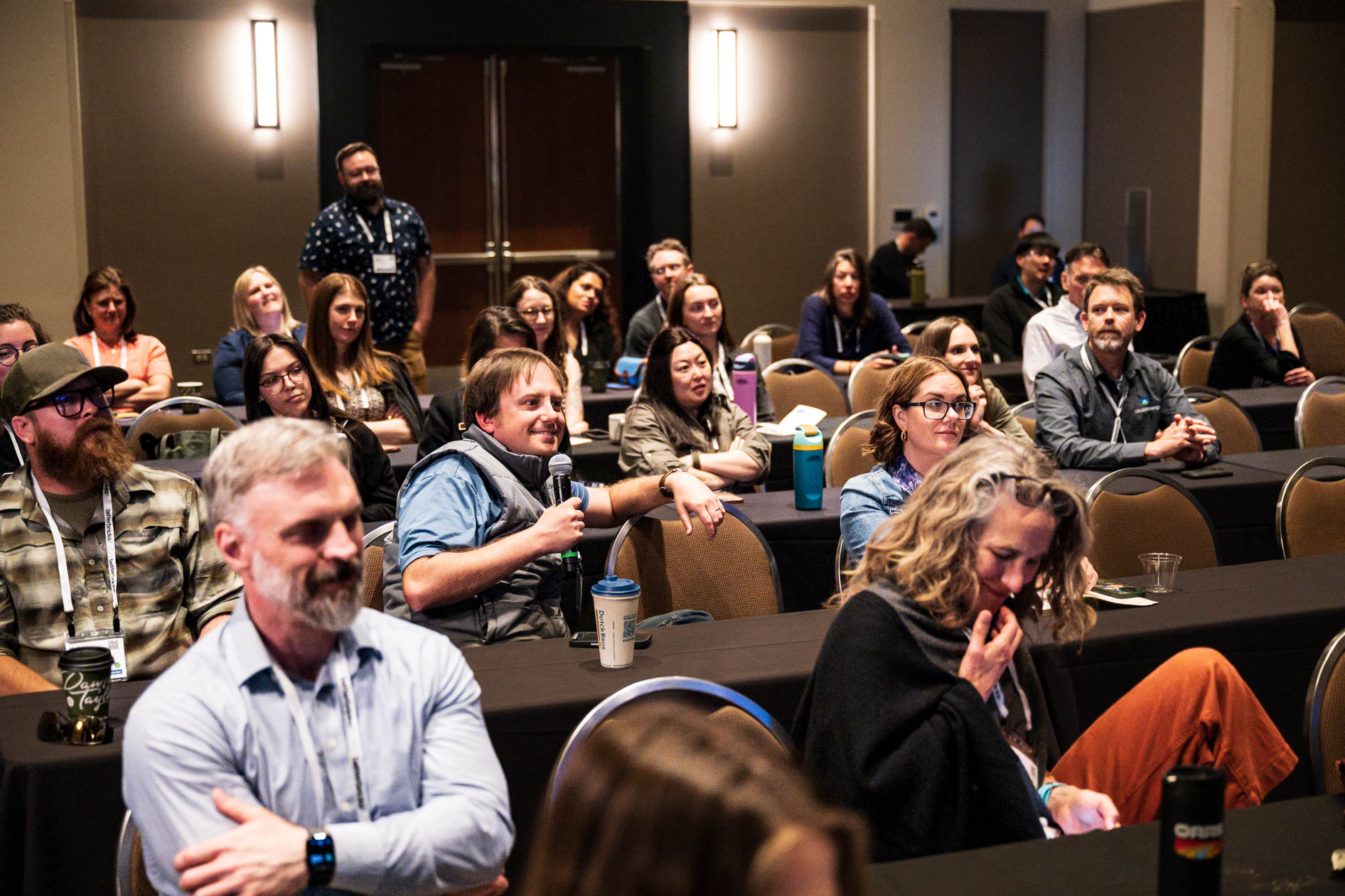 An audience member holding a microphone asks a question during a session at the Efficiency Exchange 2026 conference. Other attendees sit at long tables with black tablecloths listening and looking forward.