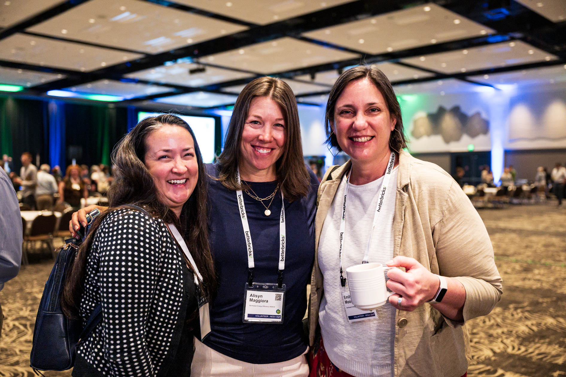 Three smiling attendees pose together inside a large convention hall during the Efficiency Exchange 2026 conference. They wear event lanyards and casual professional attire, with one holding a coffee mug.