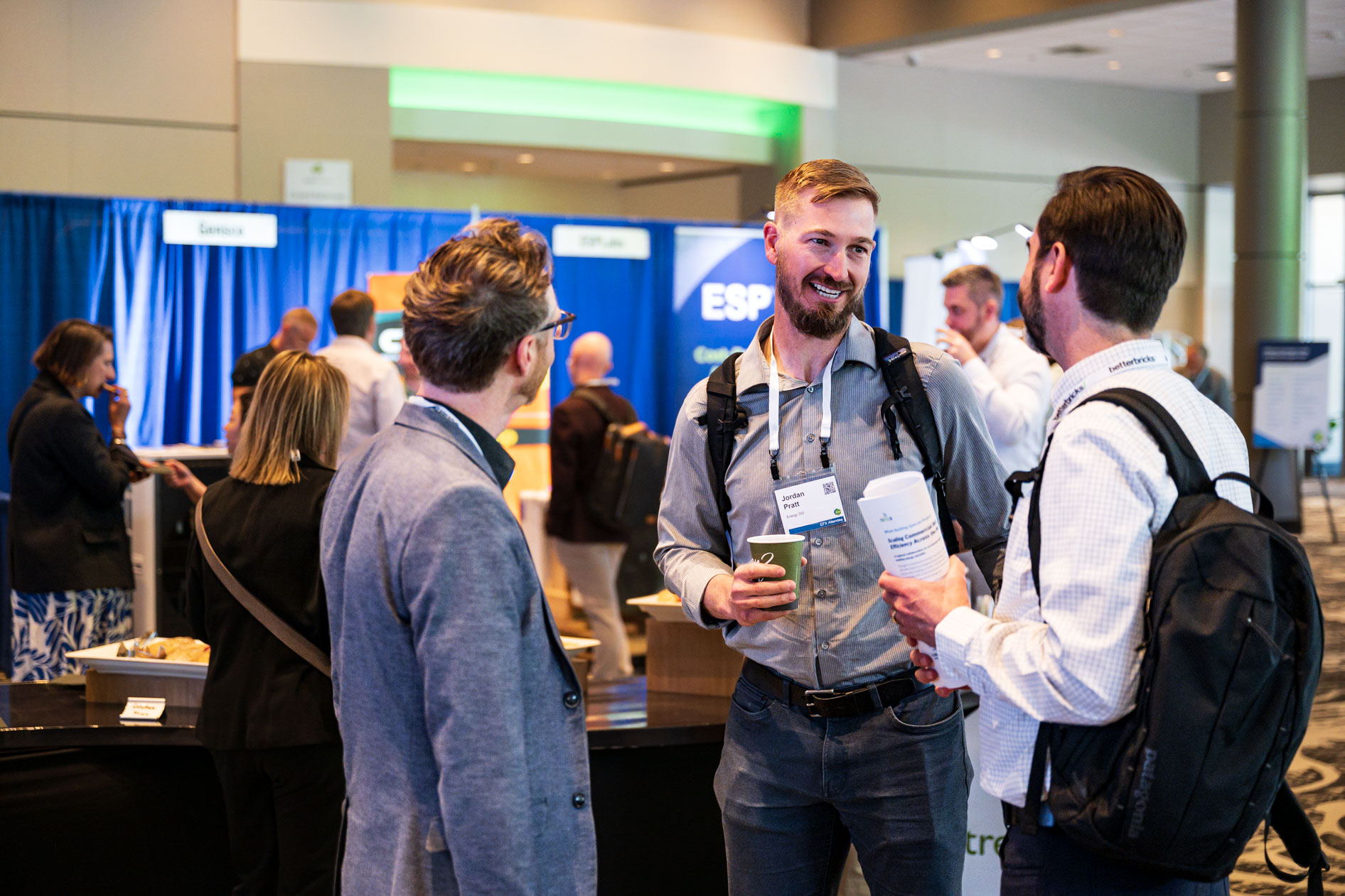 Three attendees chat and smile in the exhibition hall at the Efficiency Exchange 2026 conference. They wear event lanyards and carry professional materials, with trade show booths in the background.