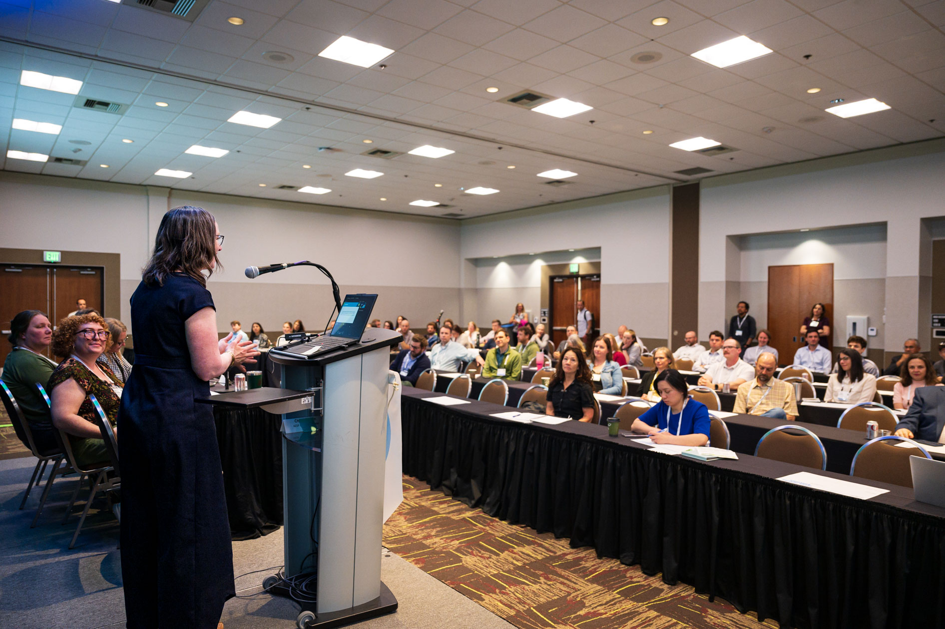 A presenter stands at a podium addressing a seated audience during a breakout session at the Efficiency Exchange 2026 conference. Attendees sit at rows of long tables with black tablecloths listening to the presentation.