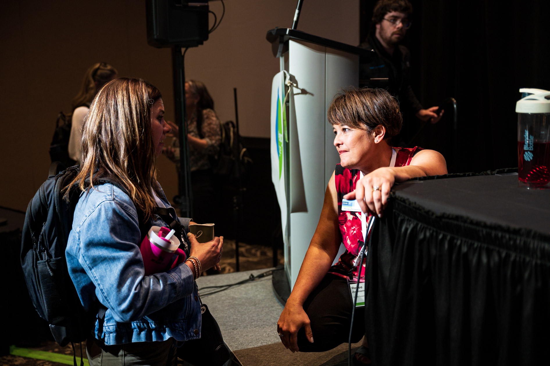 An attendee in a denim jacket chats with a speaker crouched near the stage after a session at the Efficiency Exchange 2026 conference. They converse near a podium in a dimly lit presentation hall.