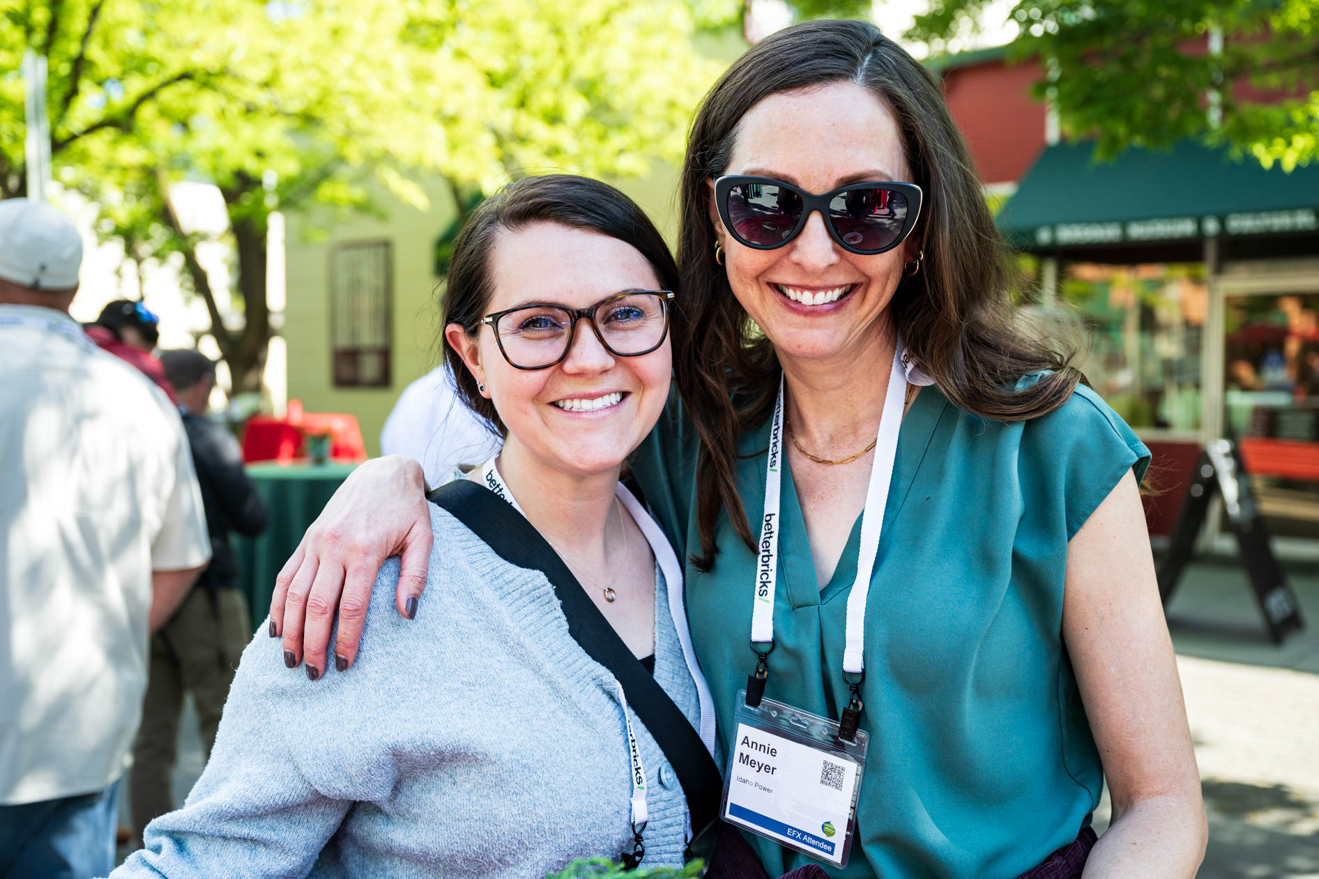 Two women smile and embrace outdoors at the Efficiency Exchange 2026 conference. They wear event lanyards and casual attire, with one wearing sunglasses and a green top, in a bright, sunlit plaza area.