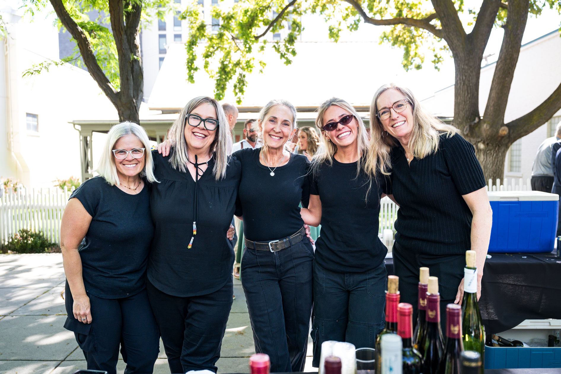 Five women wearing coordinating black outfits smile together at an outdoor reception during the Efficiency Exchange 2026 conference. They stand in front of large shade trees, with wine bottles arranged on a table in the foreground.