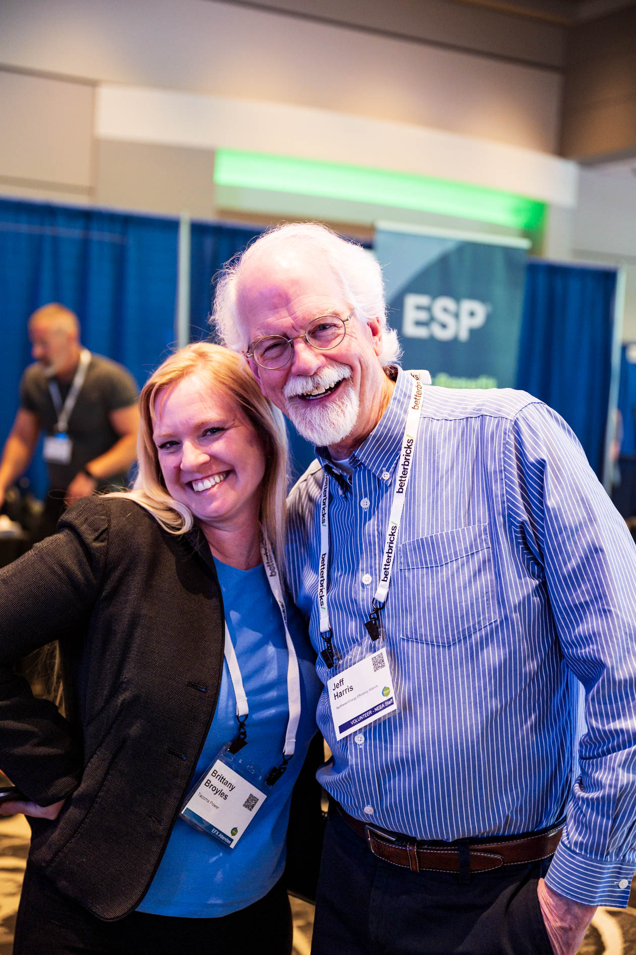 Brittany Broyles and Jeff Harris smile together inside the exhibition hall at the Efficiency Exchange 2026 conference. She wears a black blazer over a blue top, and he wears a blue striped button-down shirt, both displaying event lanyards.