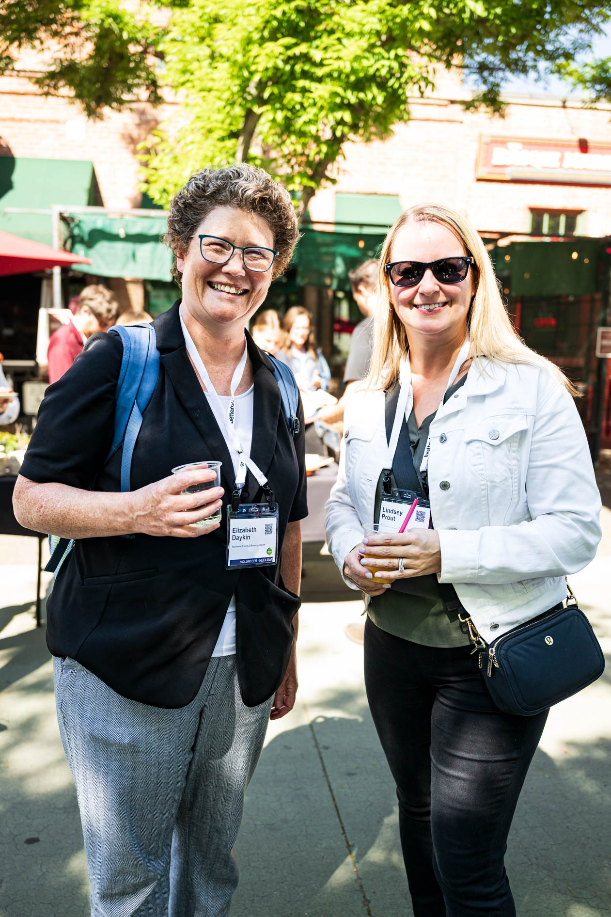 Two attendees smile outdoors at the Efficiency Exchange 2026 conference. One wears a dark blazer with glasses, and the other wears a white jacket with sunglasses, both holding beverages and wearing event name tags in a sunlit pavilion.