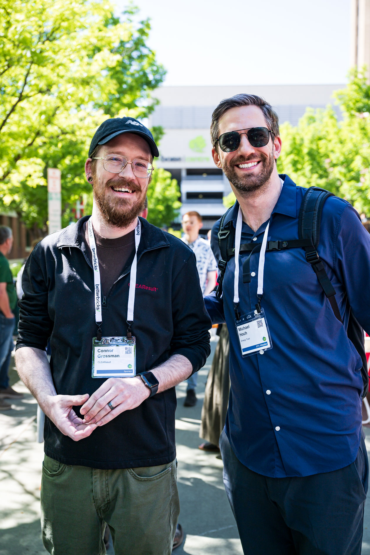 Two men smile together outdoors at the Efficiency Exchange 2026 conference. One wears a black zip-up sweater, glasses, and a baseball cap, and the other wears a blue button-down shirt and sunglasses, both wearing event badges.