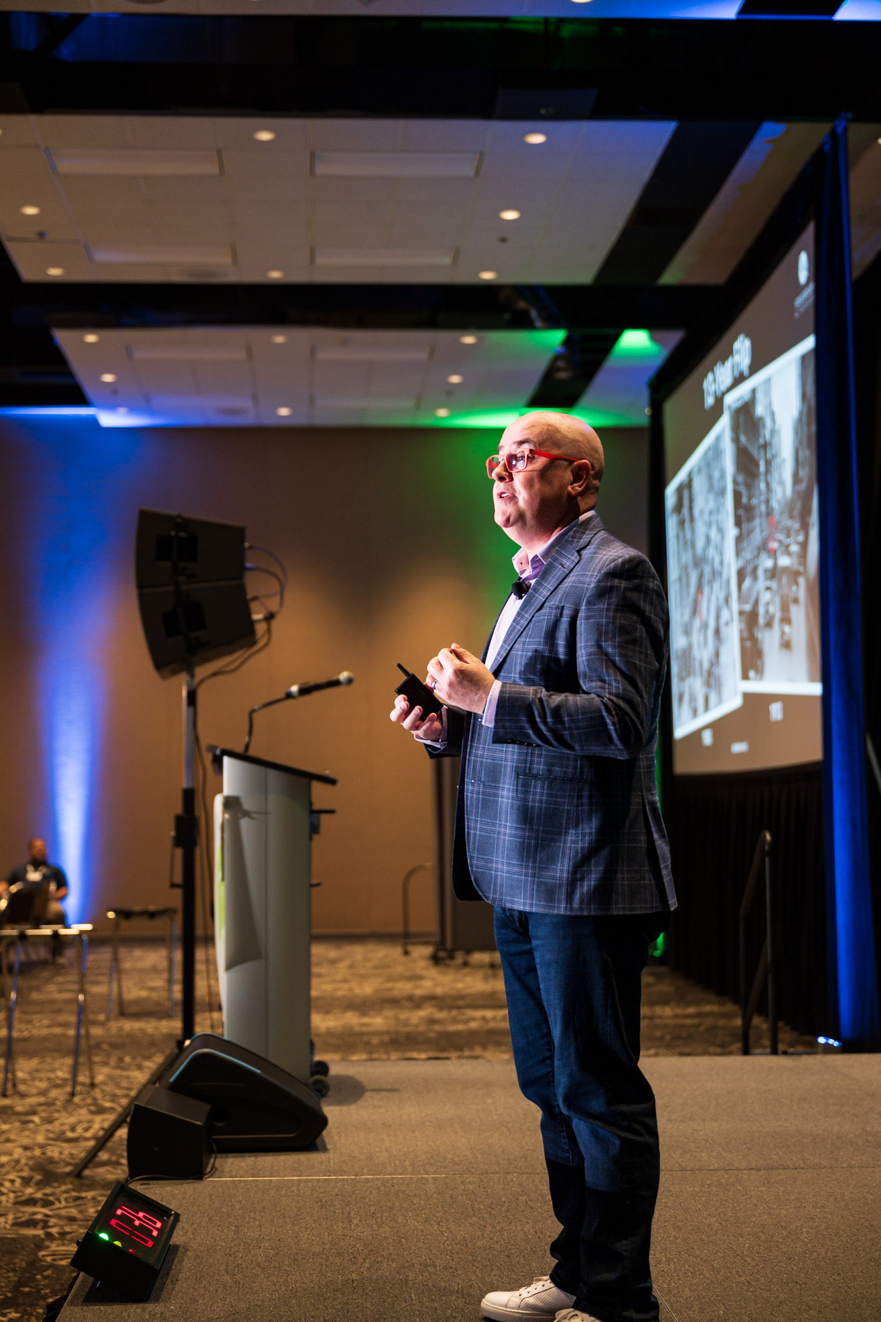 A speaker stands on stage giving a presentation at the Efficiency Exchange 2026 conference. He wears a plaid blazer, jeans, and red glasses while holding a clicker, with a presentation slide and blue and green lighting in the background.