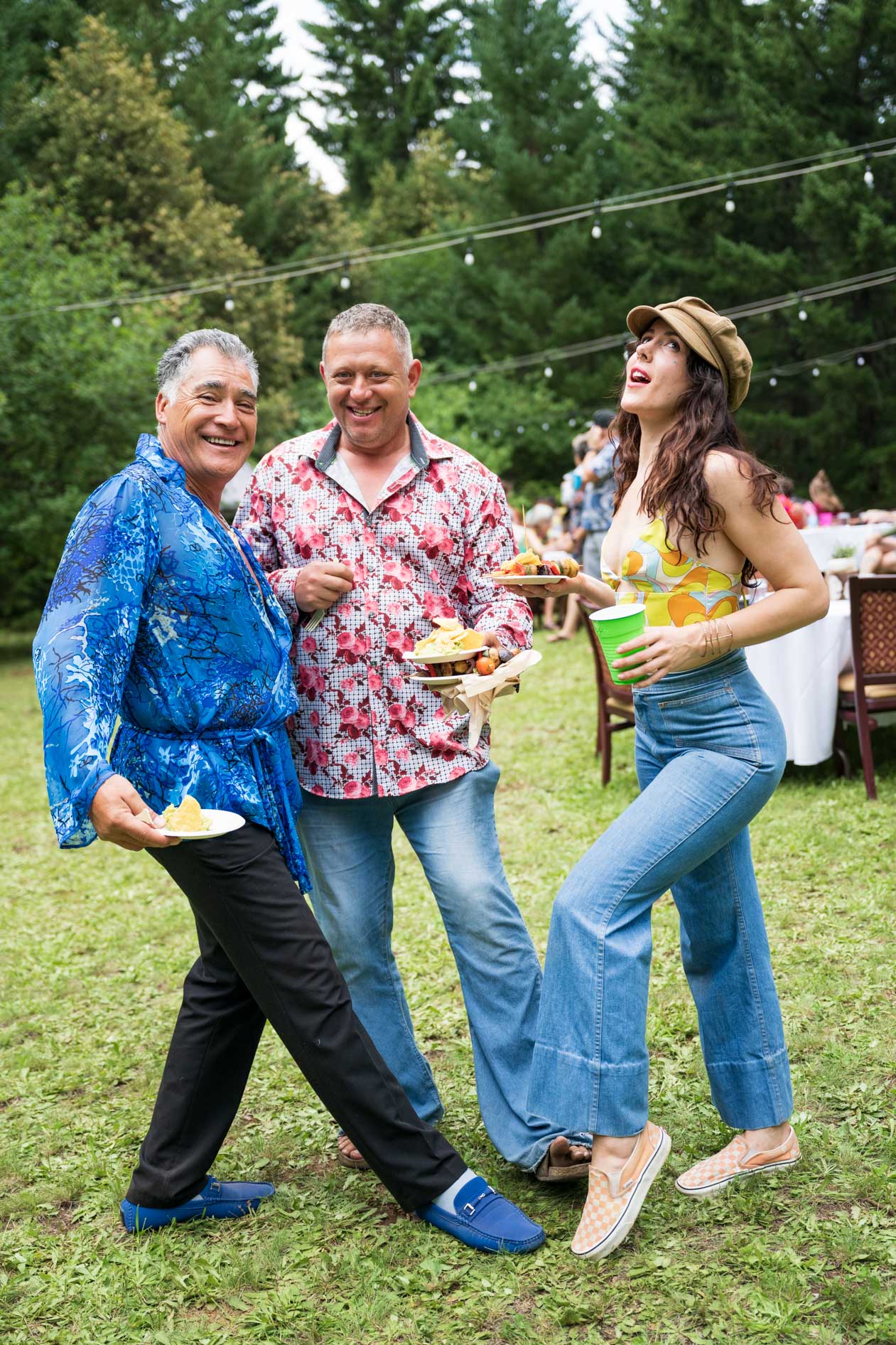Three wedding guests smiling and posing together on a green lawn at an outdoor forest wedding, showcasing expressive, colorful fashion and denim.