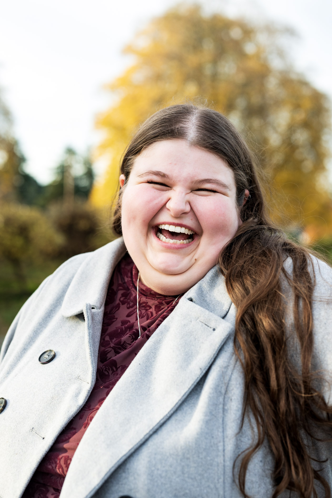 An attendee laughs joyfully for a close-up portrait outdoors during an engagement session at Peninsula Park, wearing a grey buttoned coat over a patterned maroon top.