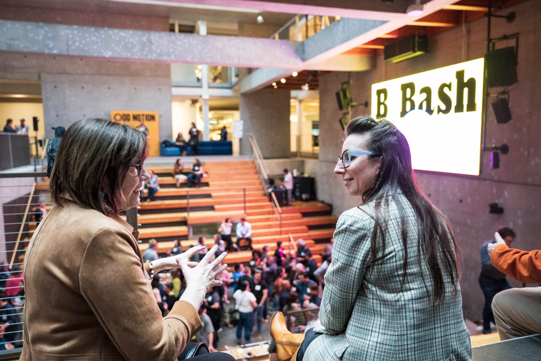 Two professionals in conversation on a balcony overlooking the B Bash event at Wieden + Kennedy. In the background, the atrium is filled with attendees and a bright "B Bash" sign, capturing the collaborative atmosphere of the Portland B Corp community.