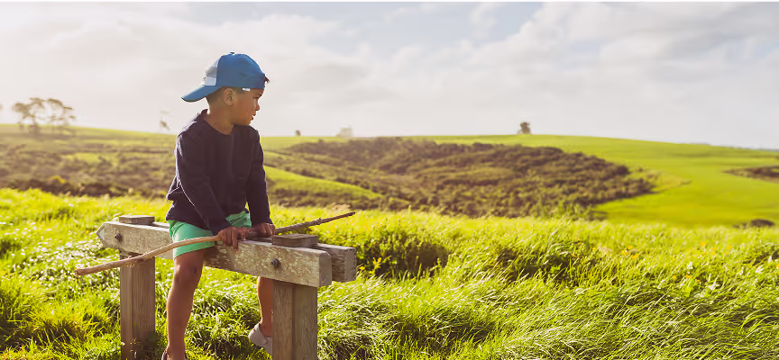 A boy in a blue hat sits on a wooden post in a grassy field.