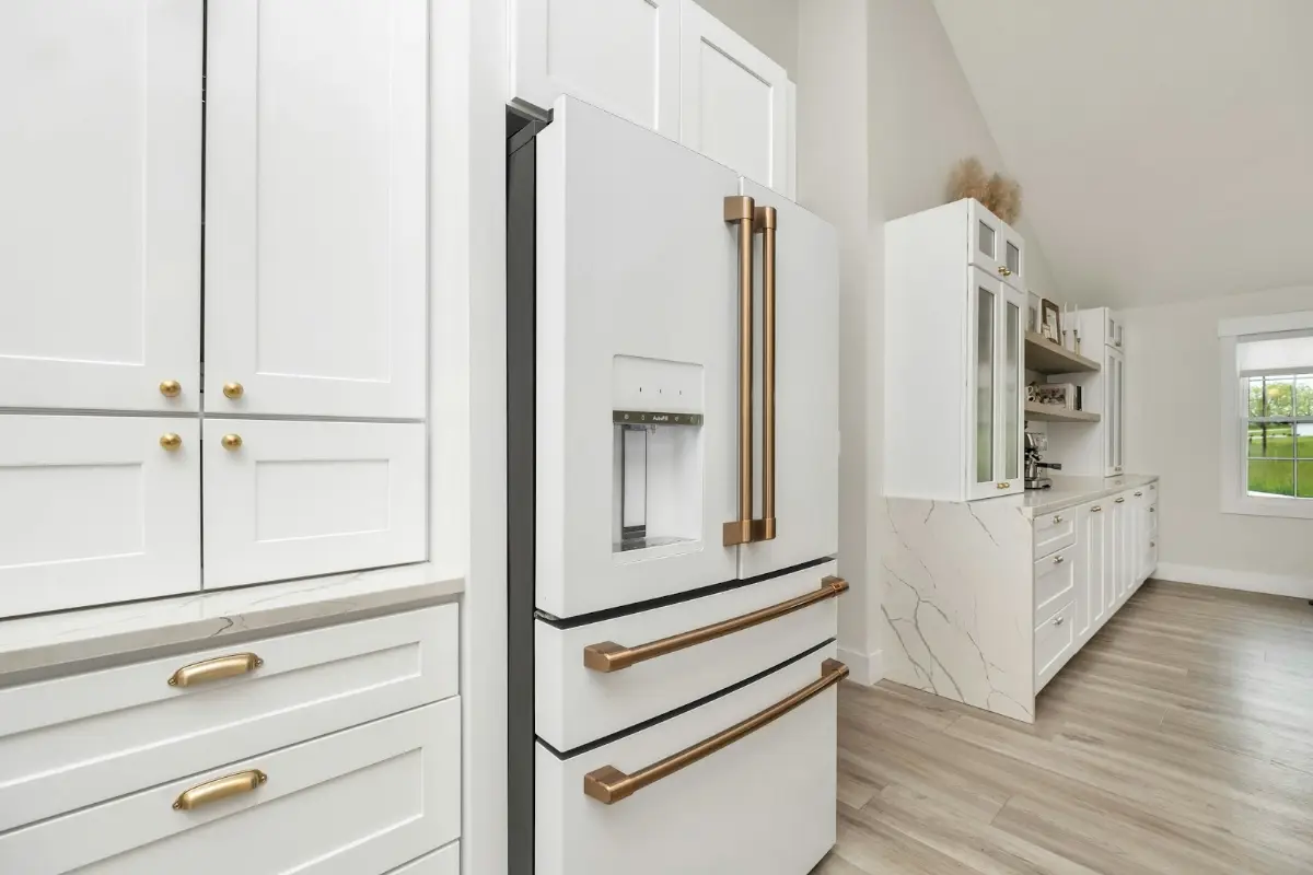 White shaker kitchen cabinets with brass pulls beside a white refrigerator in a bright remodeled kitchen